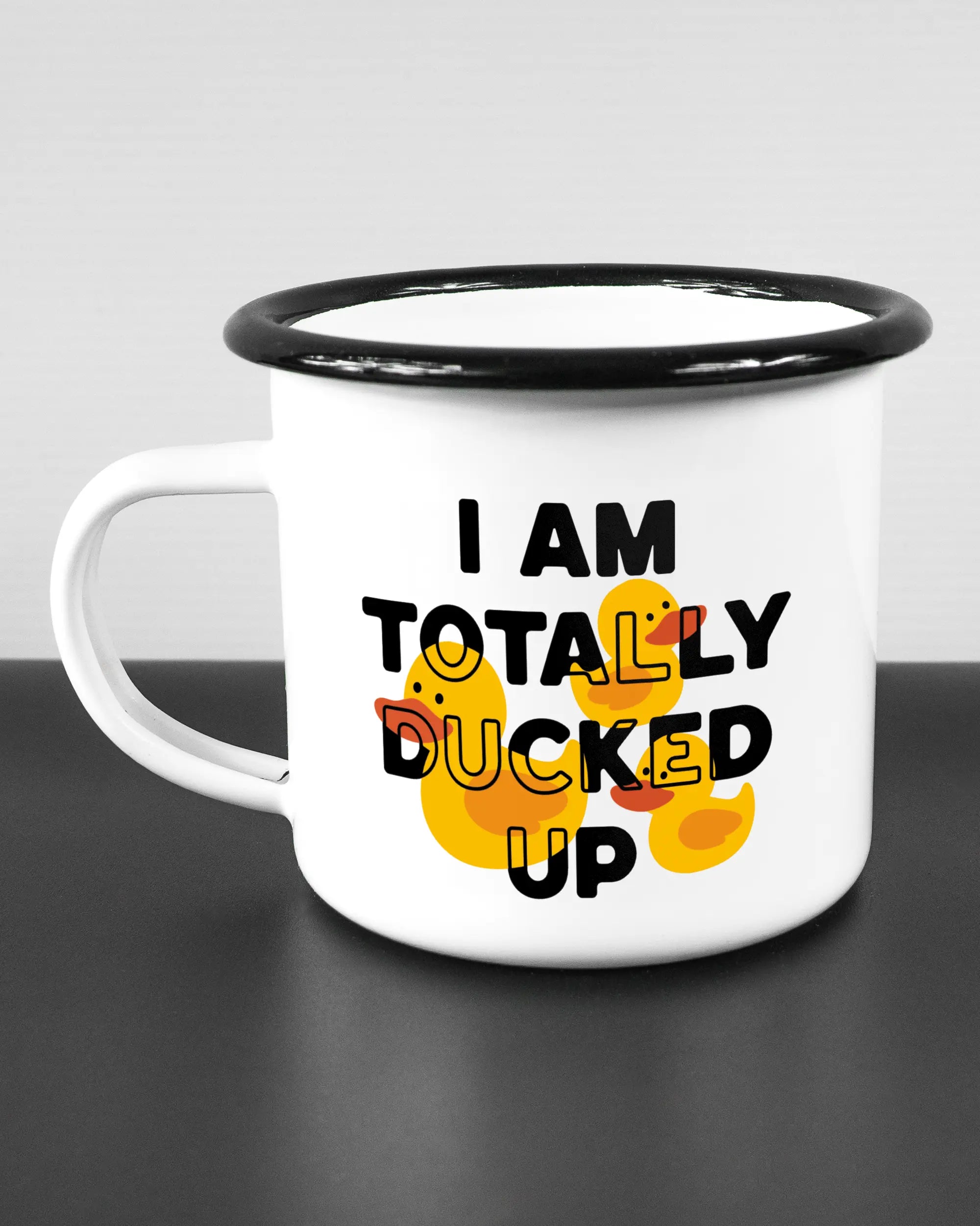 Titelbild, das die linke Seite der beidseitig bedruckten Emailletasse von Maondo mit dem Design Ducked Up in der Farbe White-Black auf einem Holztisch zeigt