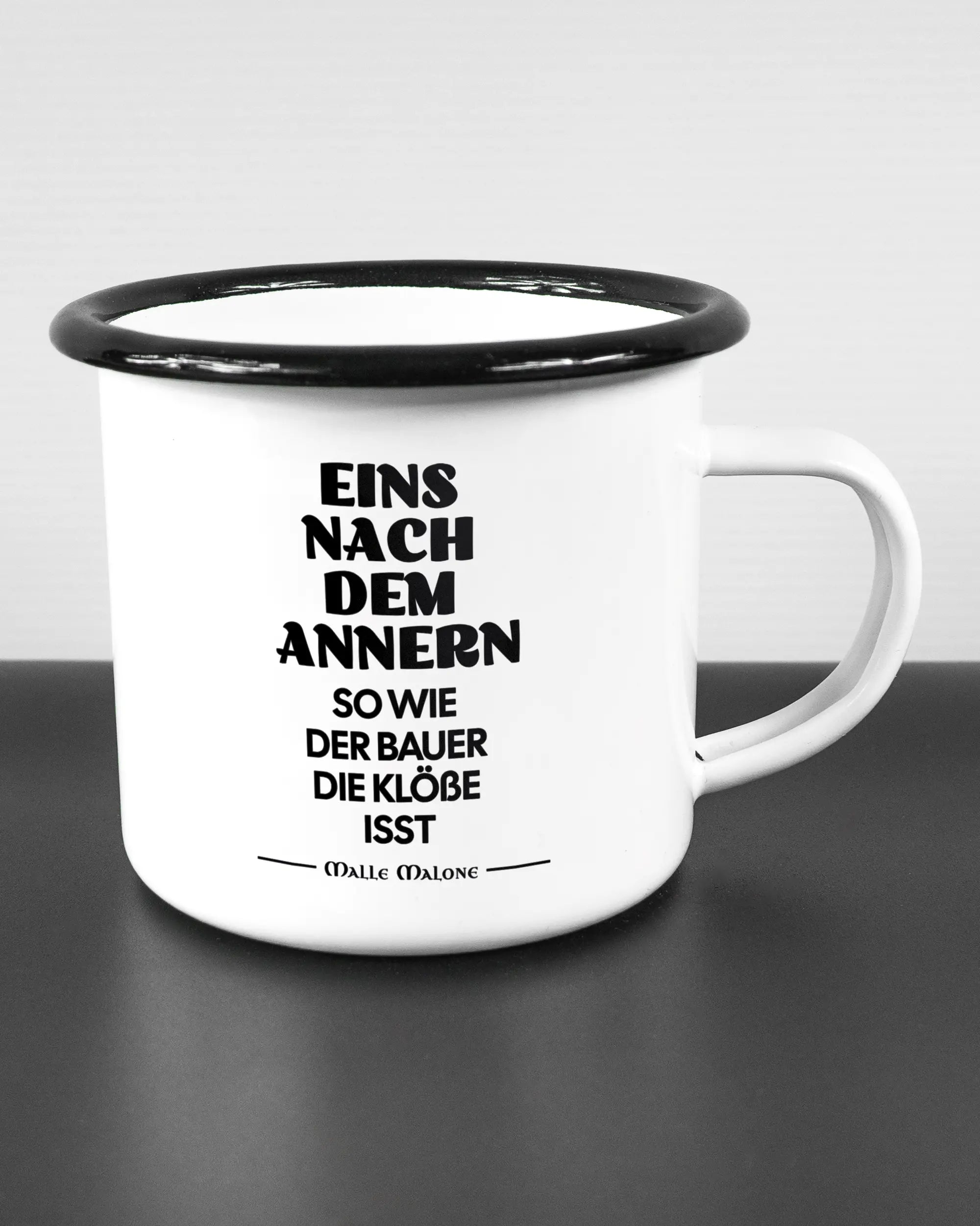 Alternativbild, das die rechte Seite der beidseitig bedruckten Emailletasse von Maondo mit dem Design Eins nach dem Annern in der Farbe White-Black auf einem Holztisch zeigt