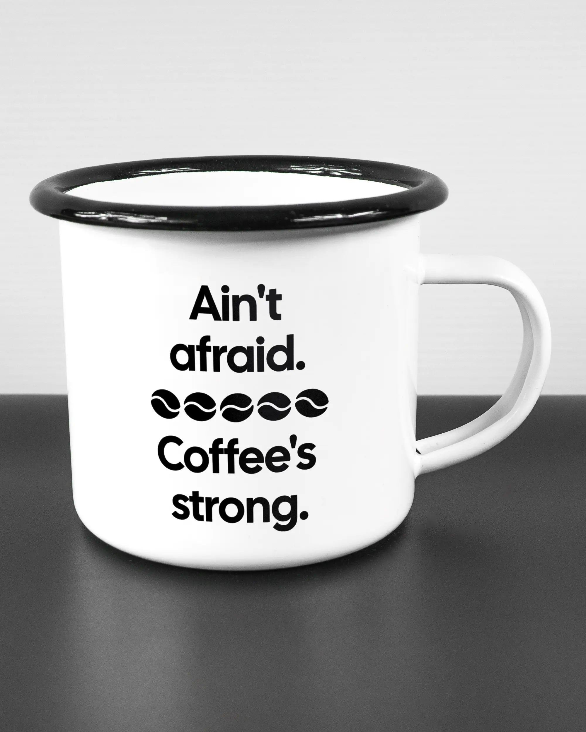 Alternativbild, das die rechte Seite der beidseitig bedruckten Emailletasse von Maondo mit dem Design Coffee is strong in der Farbe White-Black auf einem Holztisch zeigt