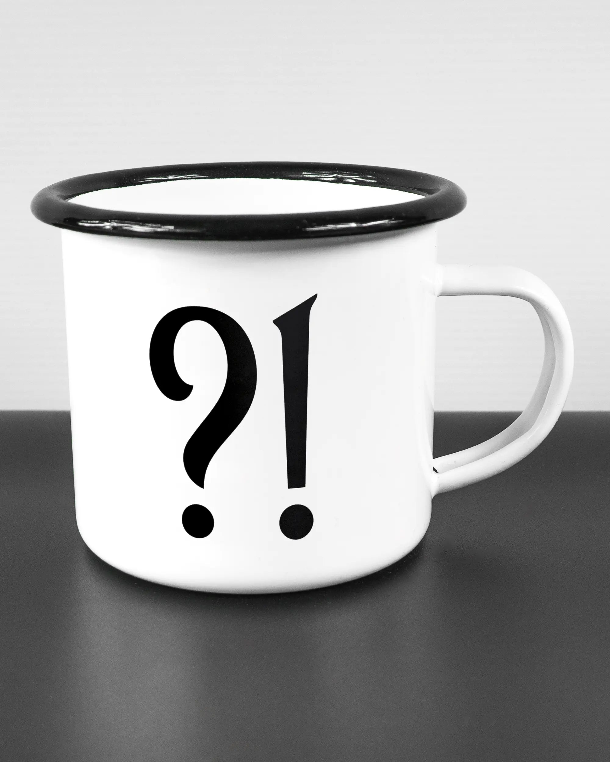 Alternativbild, das die rechte Seite der beidseitig bedruckten Emailletasse von Maondo mit dem Design WHAT in der Farbe White-Black auf einem Holztisch zeigt