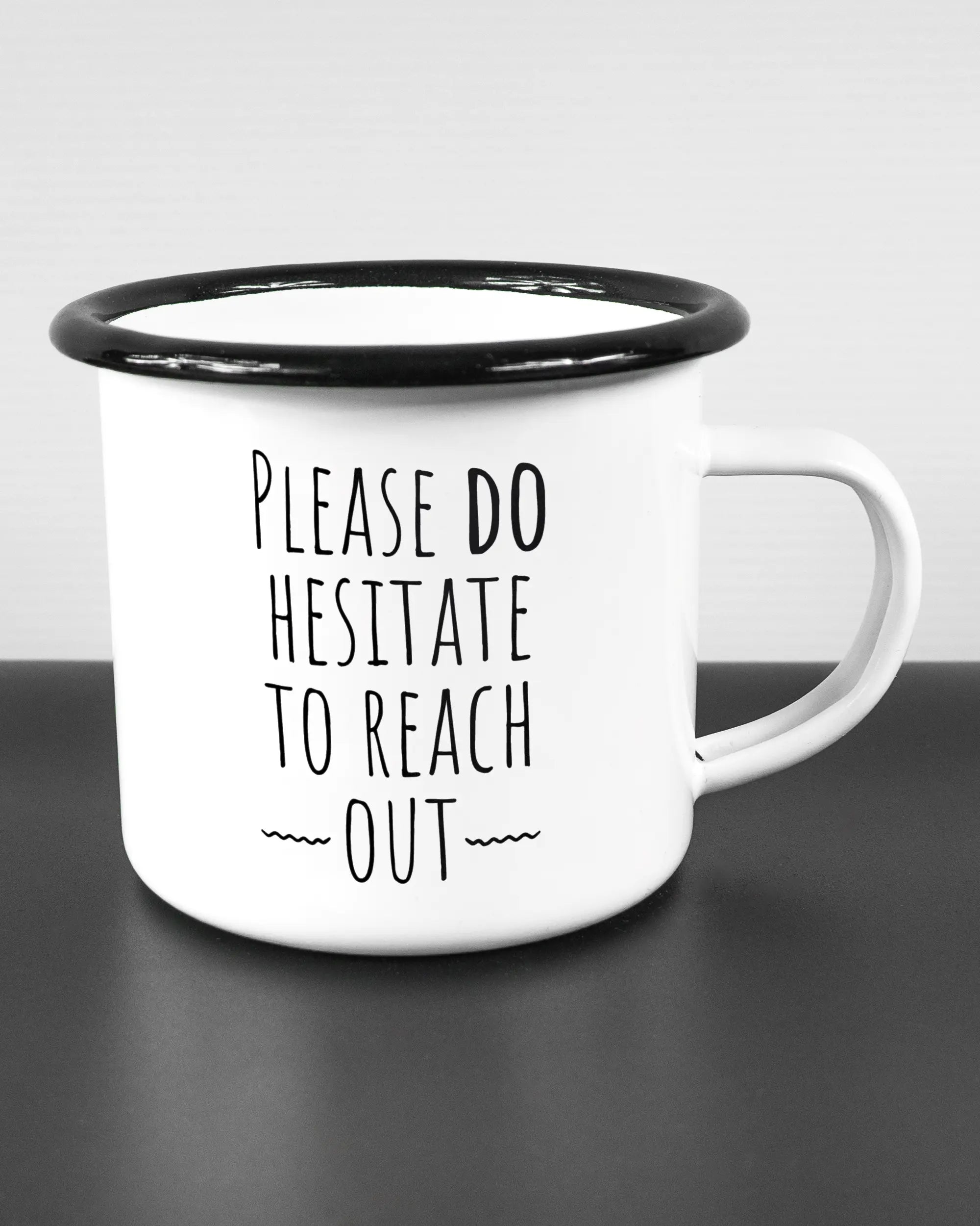 Alternativbild, das die rechte Seite der beidseitig bedruckten Emailletasse von Maondo mit dem Design Please Hesitate in der Farbe White-Black auf einem Holztisch zeigt