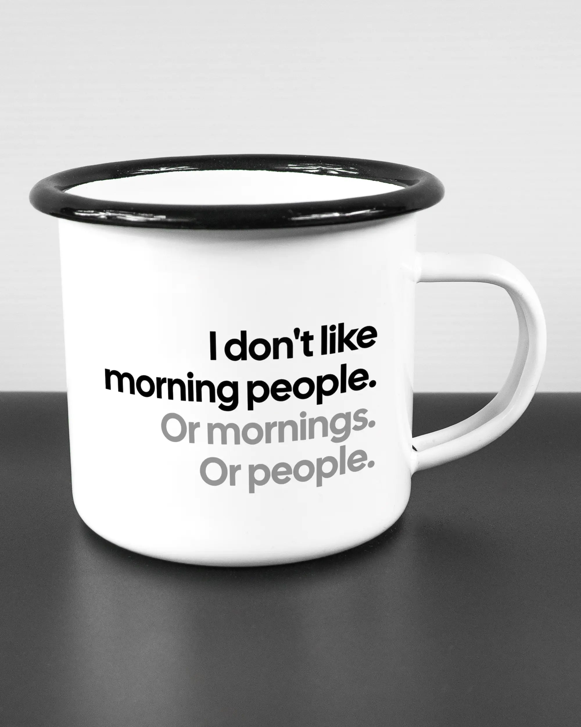 Alternativbild, das die rechte Seite der beidseitig bedruckten Emailletasse von Maondo mit dem Design Morning Person in der Farbe White-Black auf einem Holztisch zeigt