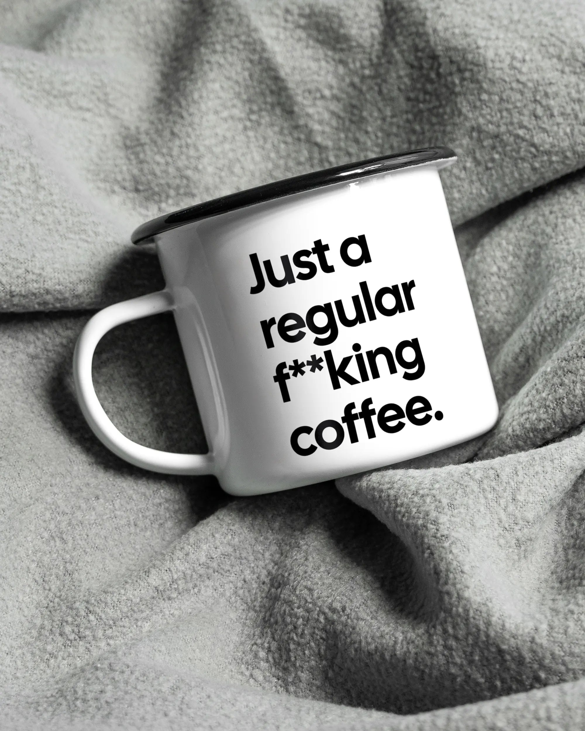 Aufnahme, die die linke Seite der beidseitig bedruckten Emailletasse von Maondo auf einer flauschigen Fließdecke mit dem Design Regular Coffee in der Farbe White-Black zeigt