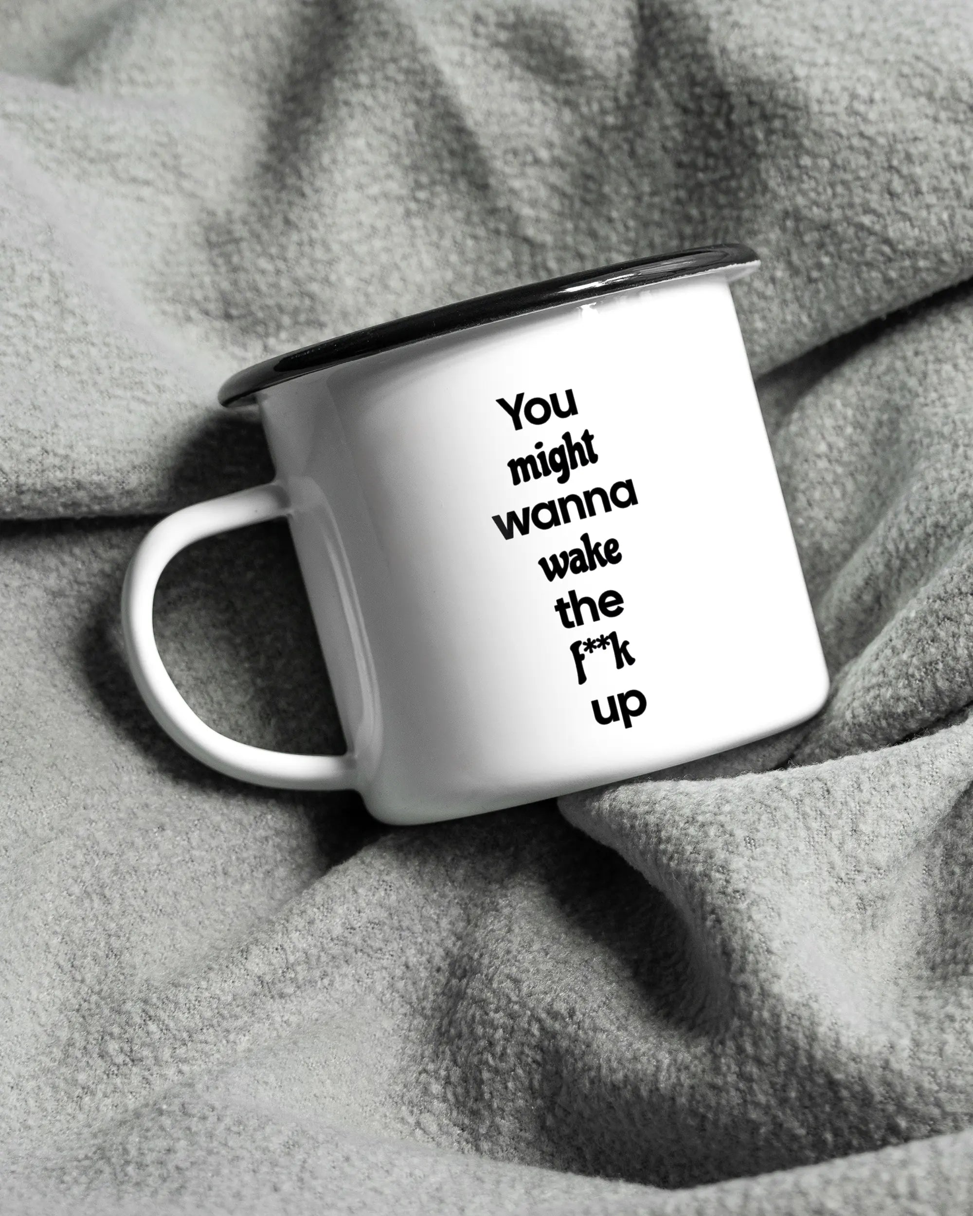 Aufnahme, die die linke Seite der beidseitig bedruckten Emailletasse von Maondo auf einer flauschigen Fließdecke mit dem Design Wanna Wake in der Farbe White-Black zeigt