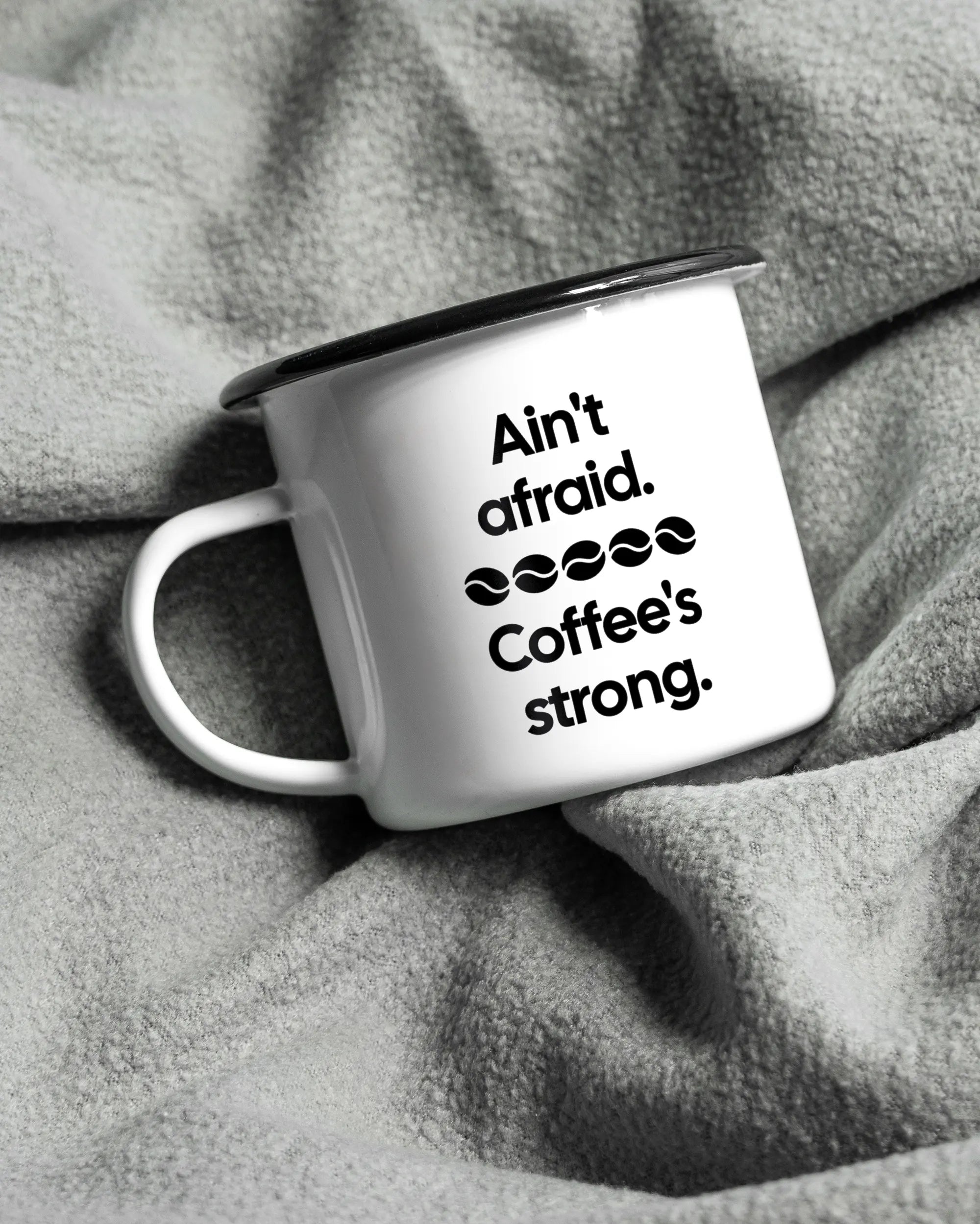 Aufnahme, die die linke Seite der beidseitig bedruckten Emailletasse von Maondo auf einer flauschigen Fließdecke mit dem Design Coffee is strong in der Farbe White-Black zeigt