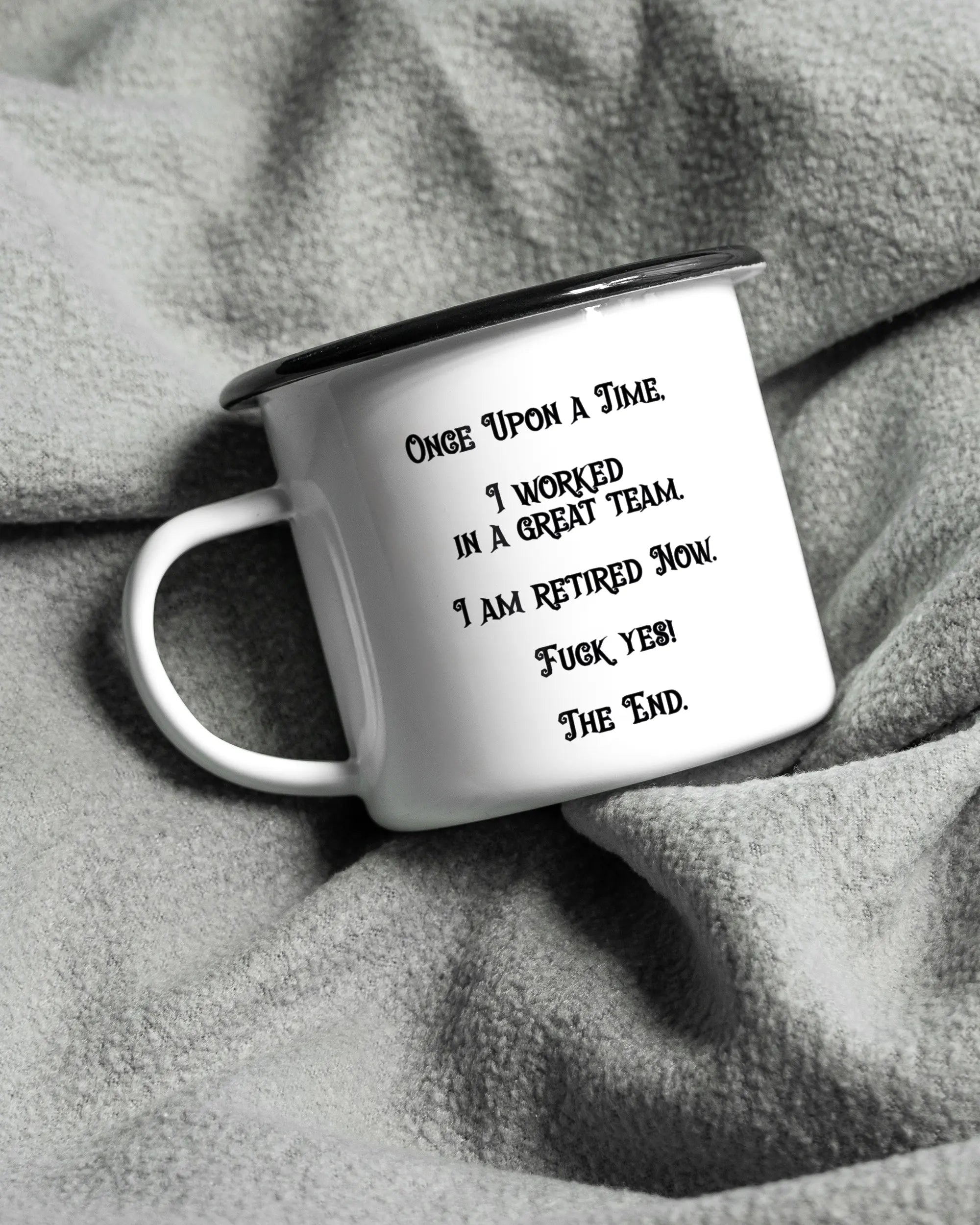 Aufnahme, die die linke Seite der beidseitig bedruckten Emailletasse von Maondo auf einer flauschigen Fließdecke mit dem Design Finally Retired in der Farbe White-Black zeigt