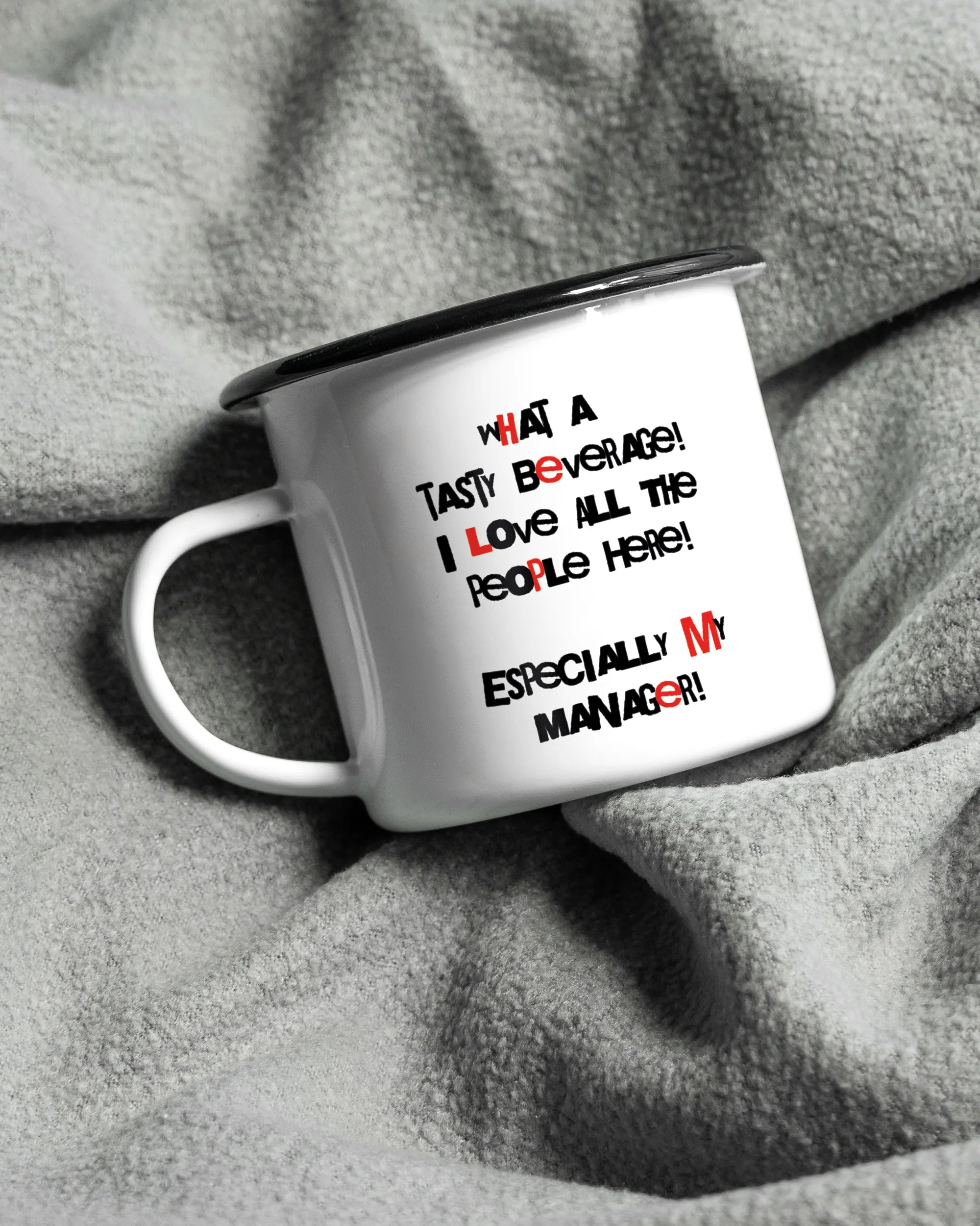 Aufnahme, die die linke Seite der beidseitig bedruckten Emailletasse von Maondo auf einer flauschigen Fließdecke mit dem Design Help me in der Farbe White-Black zeigt