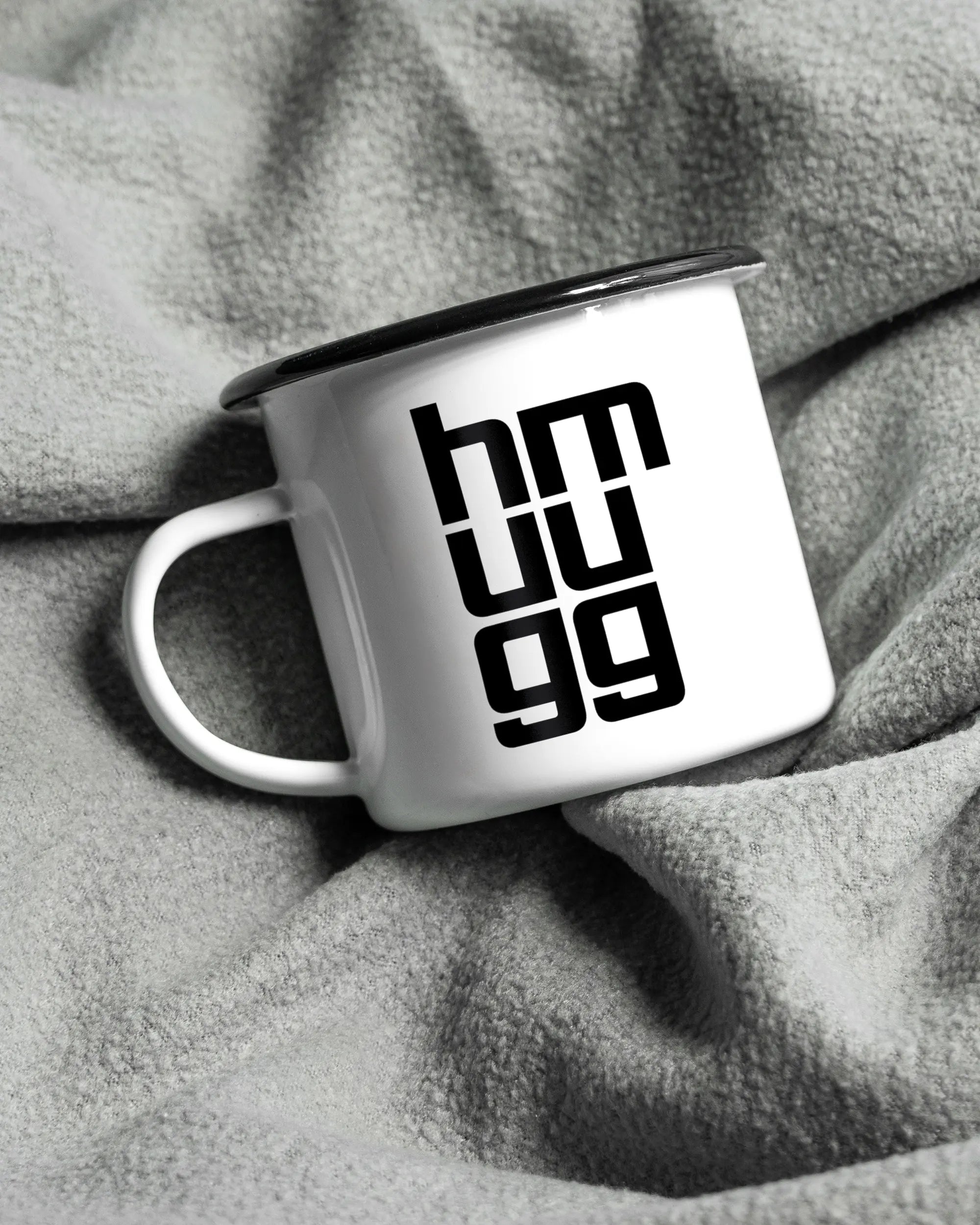 Aufnahme, die die linke Seite der beidseitig bedruckten Emailletasse von Maondo auf einer flauschigen Fließdecke mit dem Design Hug Mug in der Farbe White-Black zeigt
