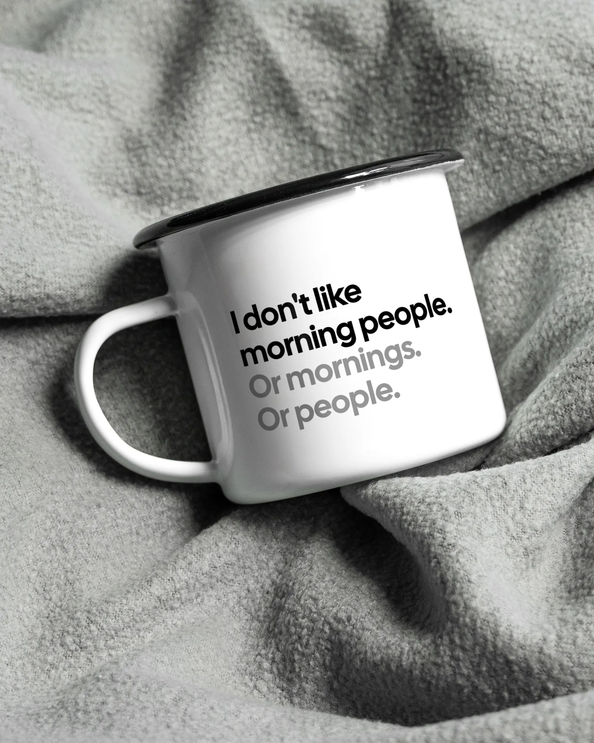 Aufnahme, die die linke Seite der beidseitig bedruckten Emailletasse von Maondo auf einer flauschigen Fließdecke mit dem Design Morning Person in der Farbe White-Black zeigt
