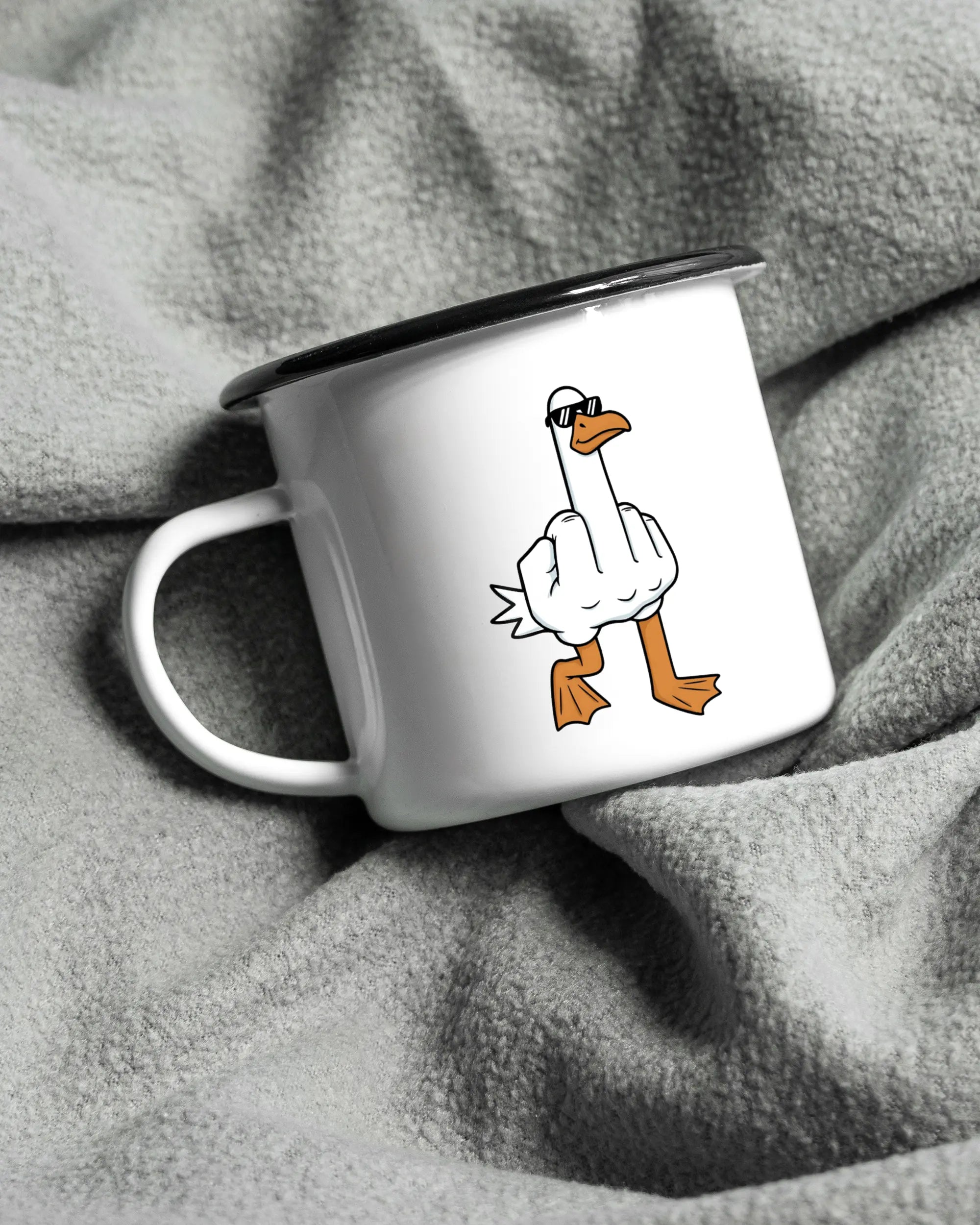 Aufnahme, die die linke Seite der beidseitig bedruckten Emailletasse von Maondo auf einer flauschigen Fließdecke mit dem Design I give a duck in der Farbe White-Black zeigt