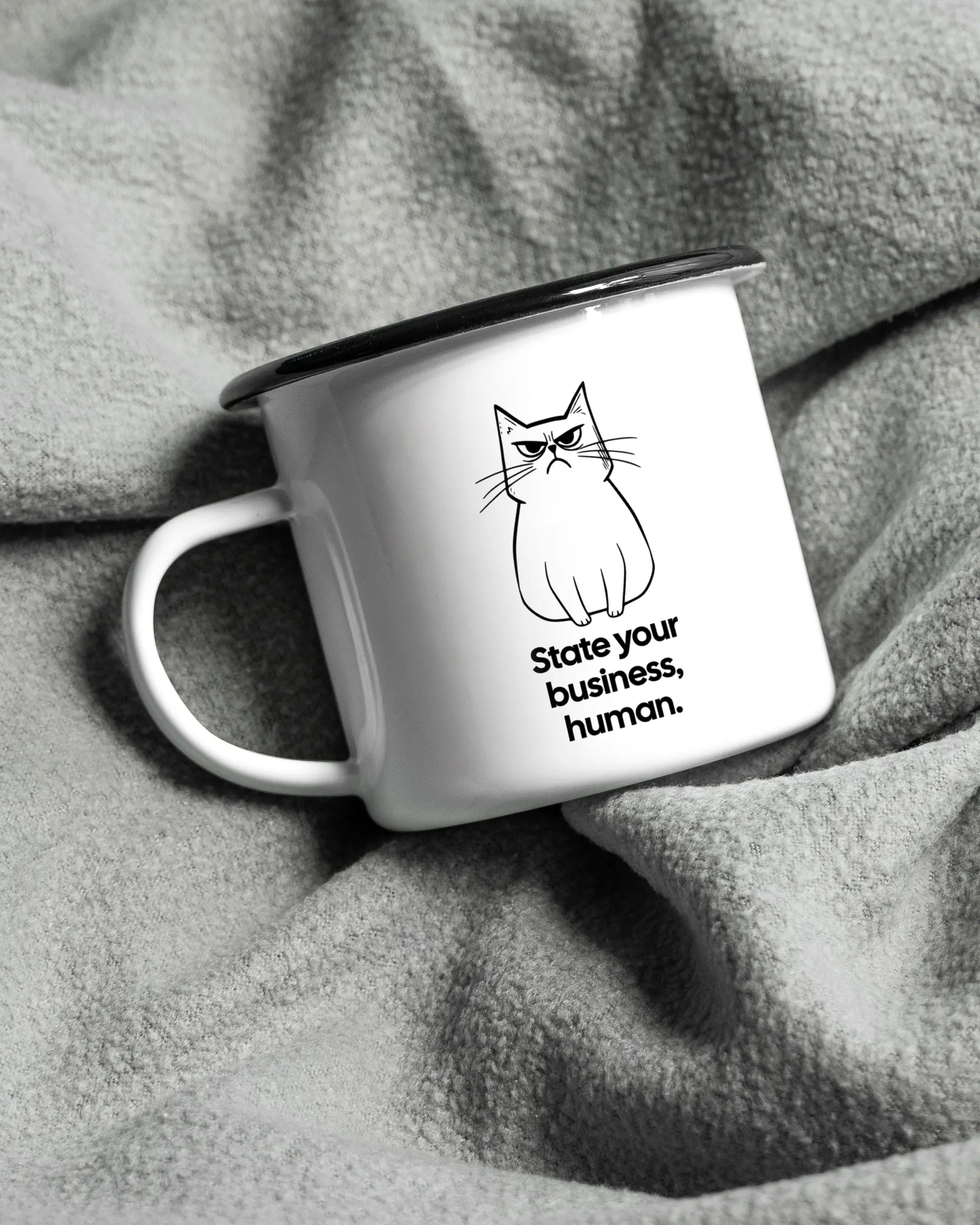 Aufnahme, die die linke Seite der beidseitig bedruckten Emailletasse von Maondo auf einer flauschigen Fließdecke mit dem Design Grumpy Purr in der Farbe White-Black zeigt