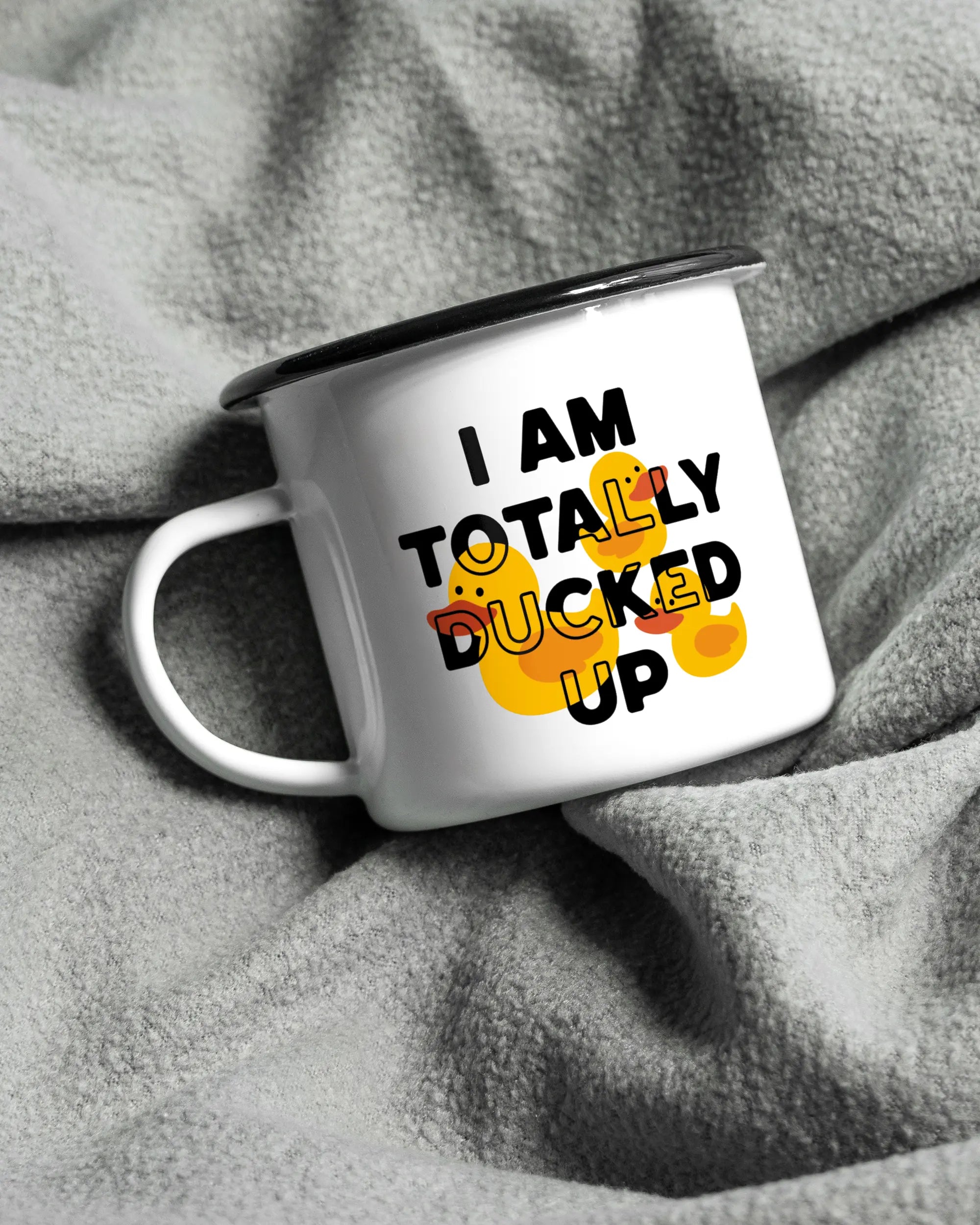 Aufnahme, die die linke Seite der beidseitig bedruckten Emailletasse von Maondo auf einer flauschigen Fließdecke mit dem Design Ducked Up in der Farbe White-Black zeigt