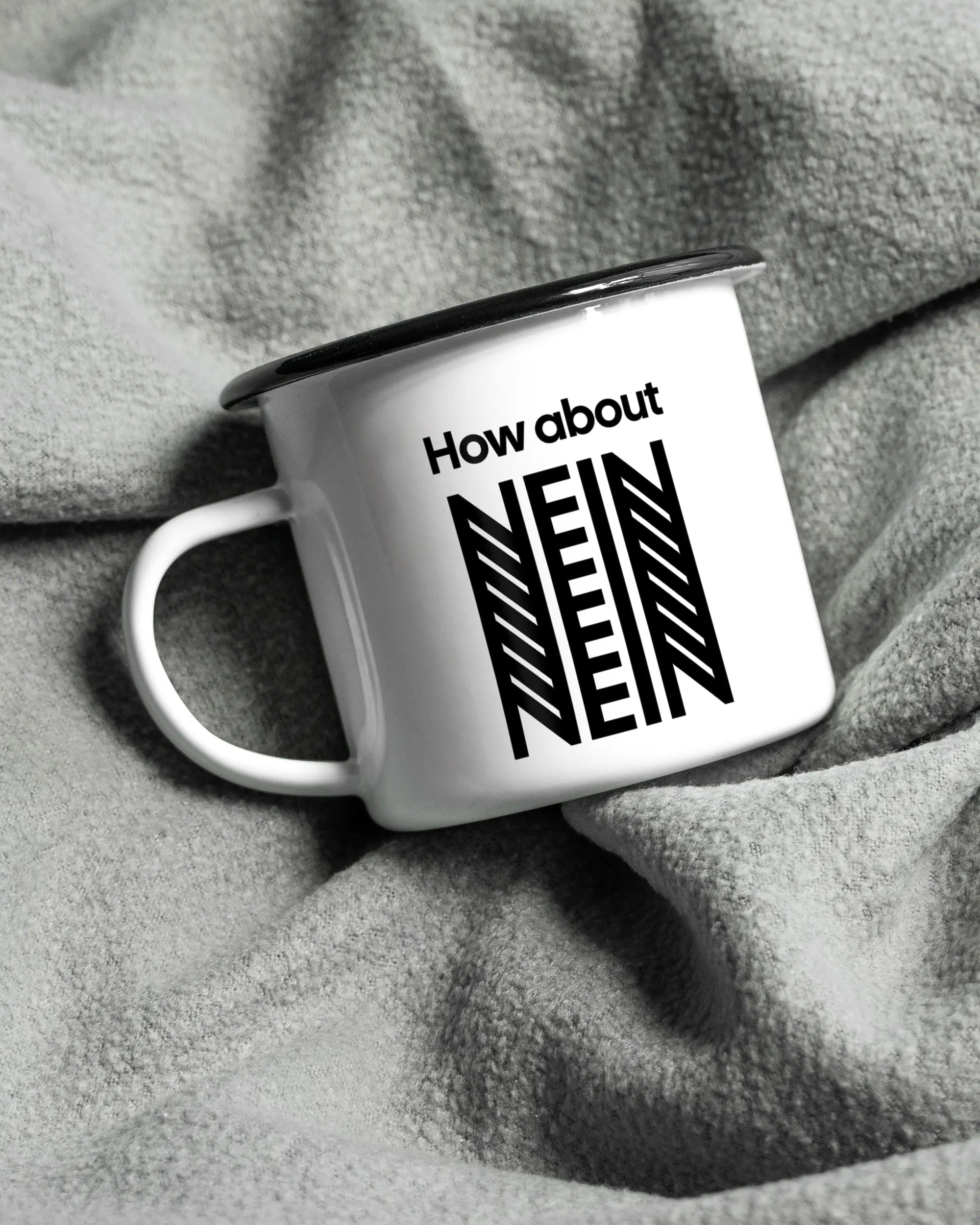 Aufnahme, die die linke Seite der beidseitig bedruckten Emailletasse von Maondo auf einer flauschigen Fließdecke mit dem Design How about nein in der Farbe White-Black zeigt