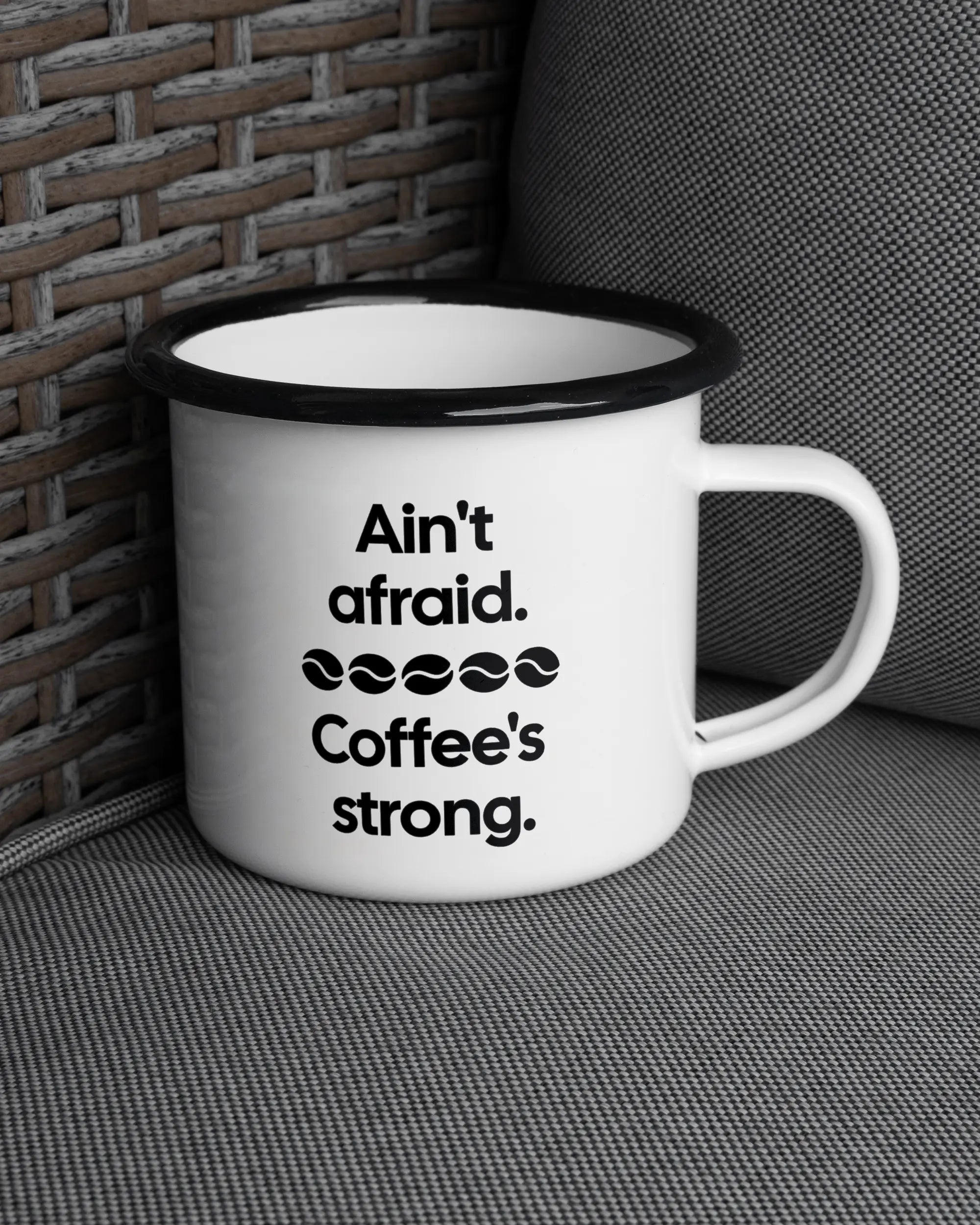 Ansicht der rechten Seite der beidseitig bedruckten Emailletasse von Maondo auf einem Balkonmöbel bzw. Campingstuhl mit dem Design Coffee is strong in der Farbe White-Black