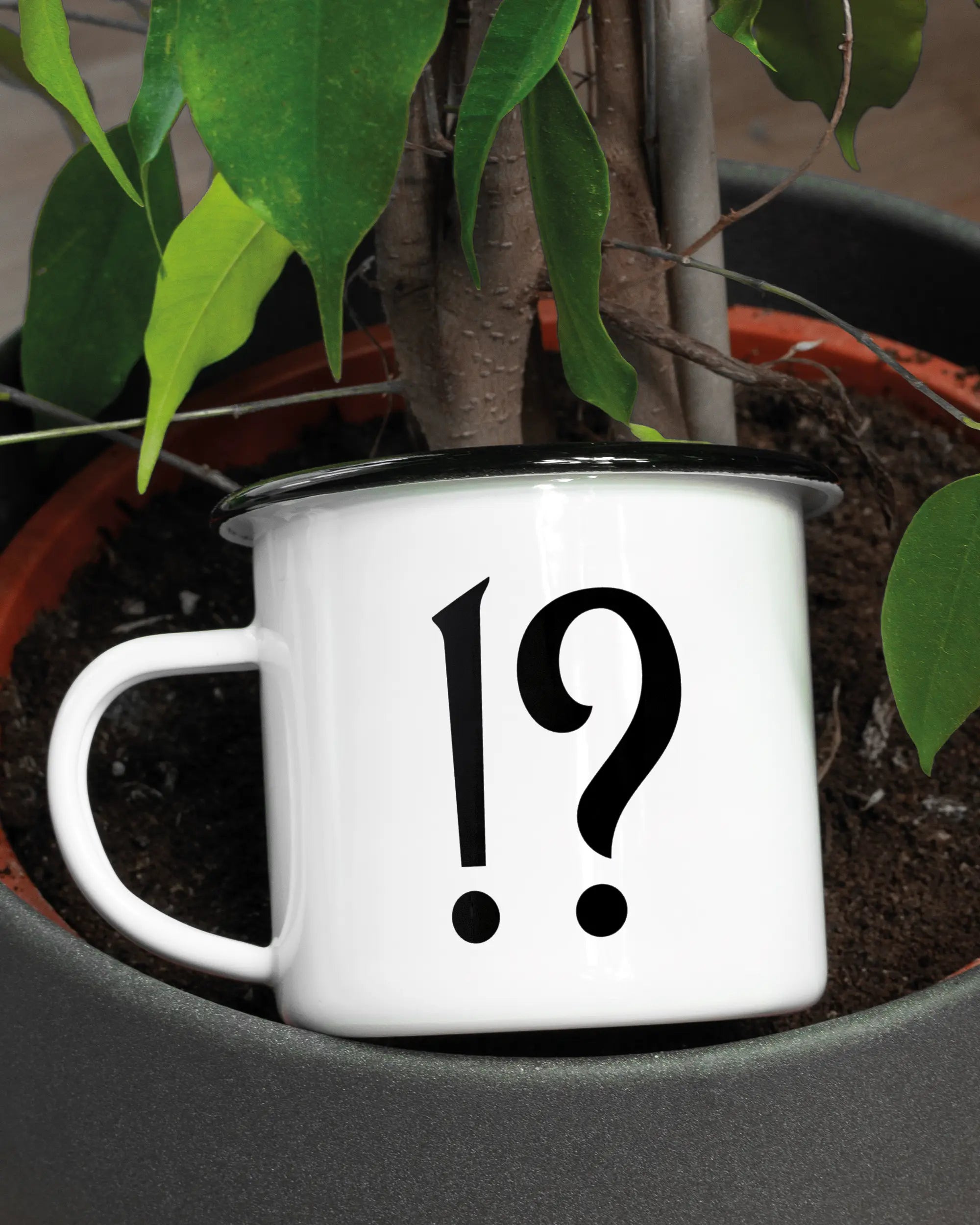 Aufnahme der linken Seite der beidseitig bedruckten Emailletasse von Maondo mit dem Design WHAT in der Farbe White-Black in einem Blumentopf