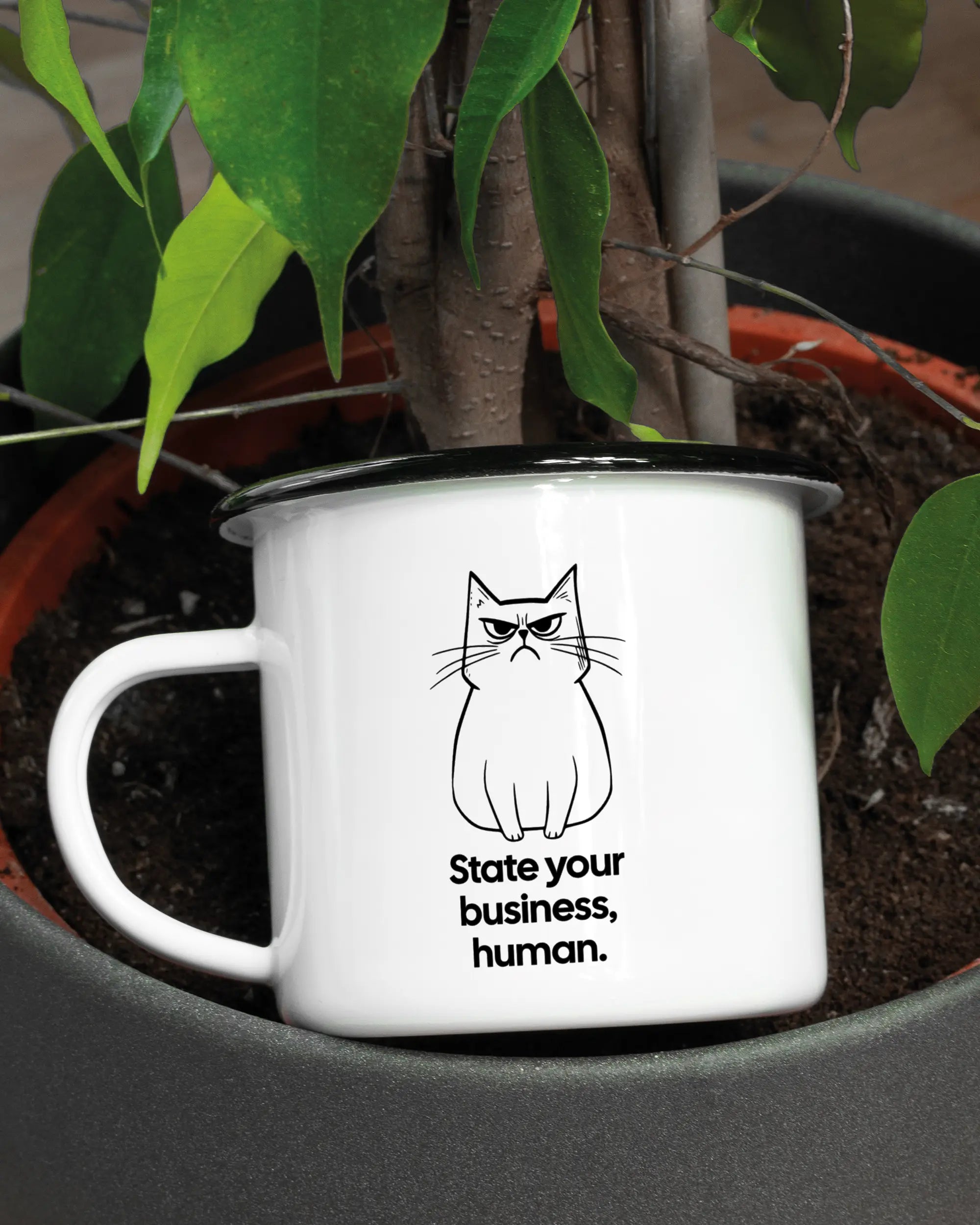 Aufnahme der linken Seite der beidseitig bedruckten Emailletasse von Maondo mit dem Design Grumpy Purr in der Farbe White-Black in einem Blumentopf