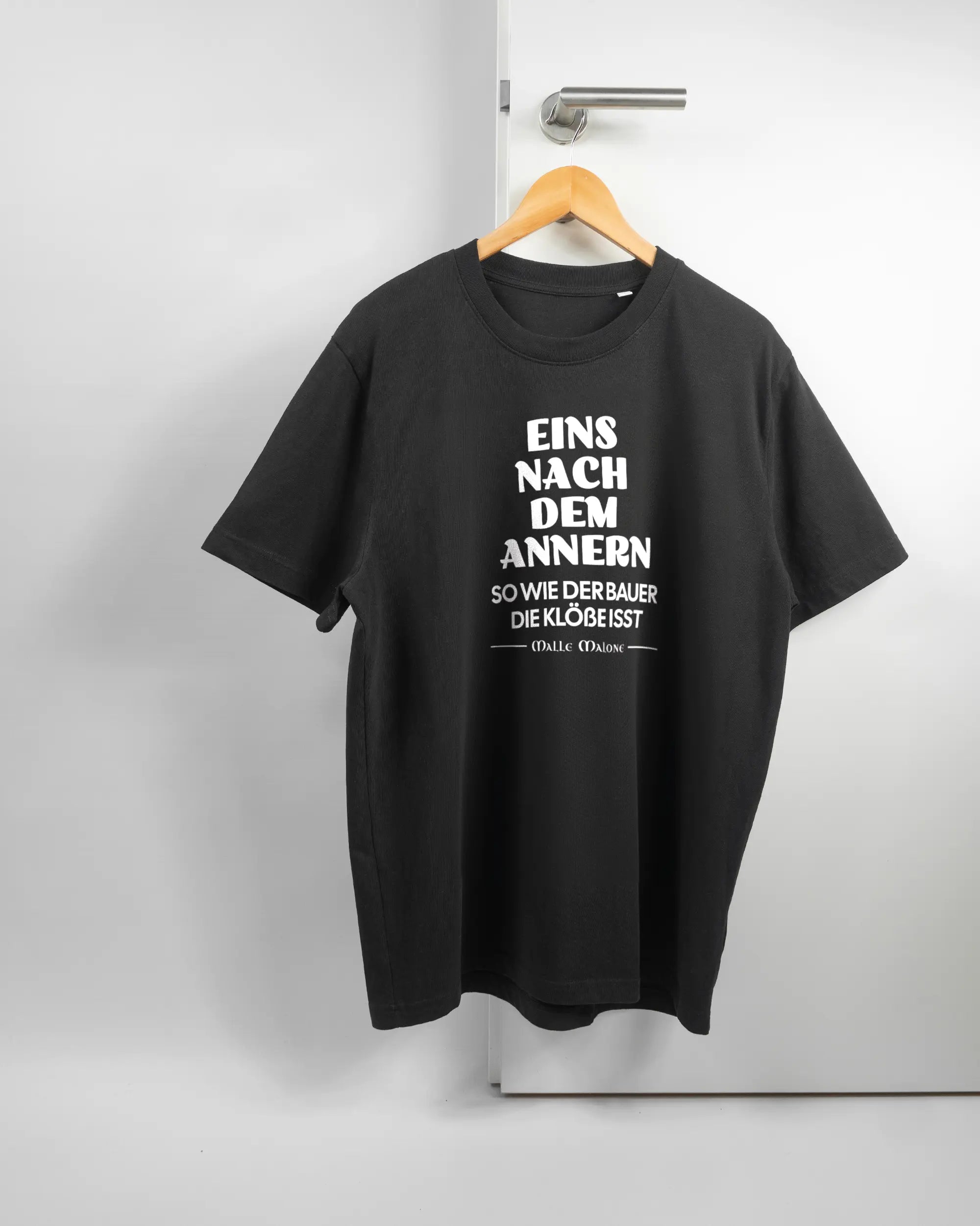 Vorderseite des Organic Unisex T-Shirt Relaxed von Maondo mit dem Design Eins nach dem Annern in der Farbe Black und in der Größe L auf einem Bügel an der Türklinke aufgehängt