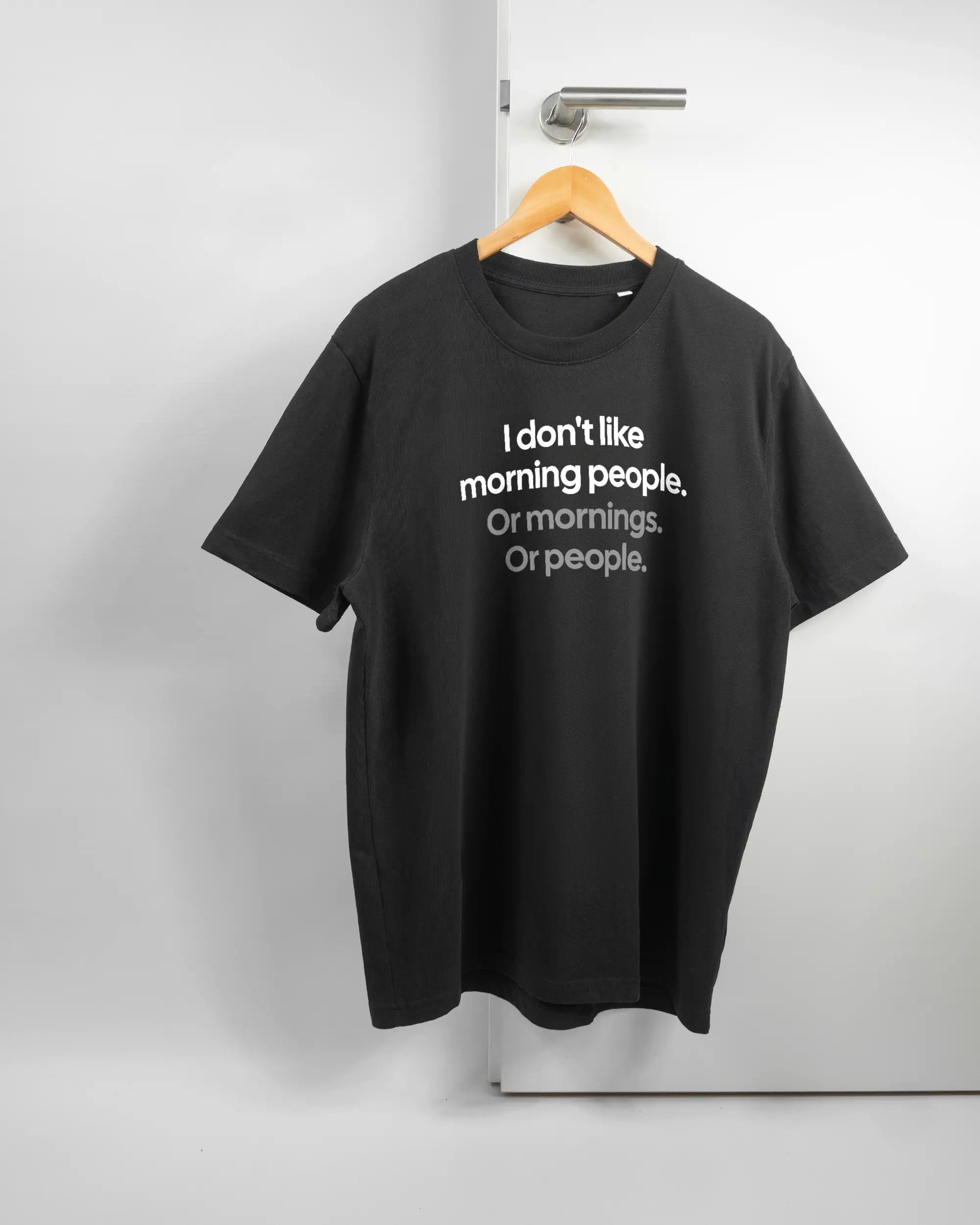 Vorderseite des Organic Unisex T-Shirt Relaxed von Maondo mit dem Design Morning Person in der Farbe Black und in der Größe L auf einem Bügel an der Türklinke aufgehängt