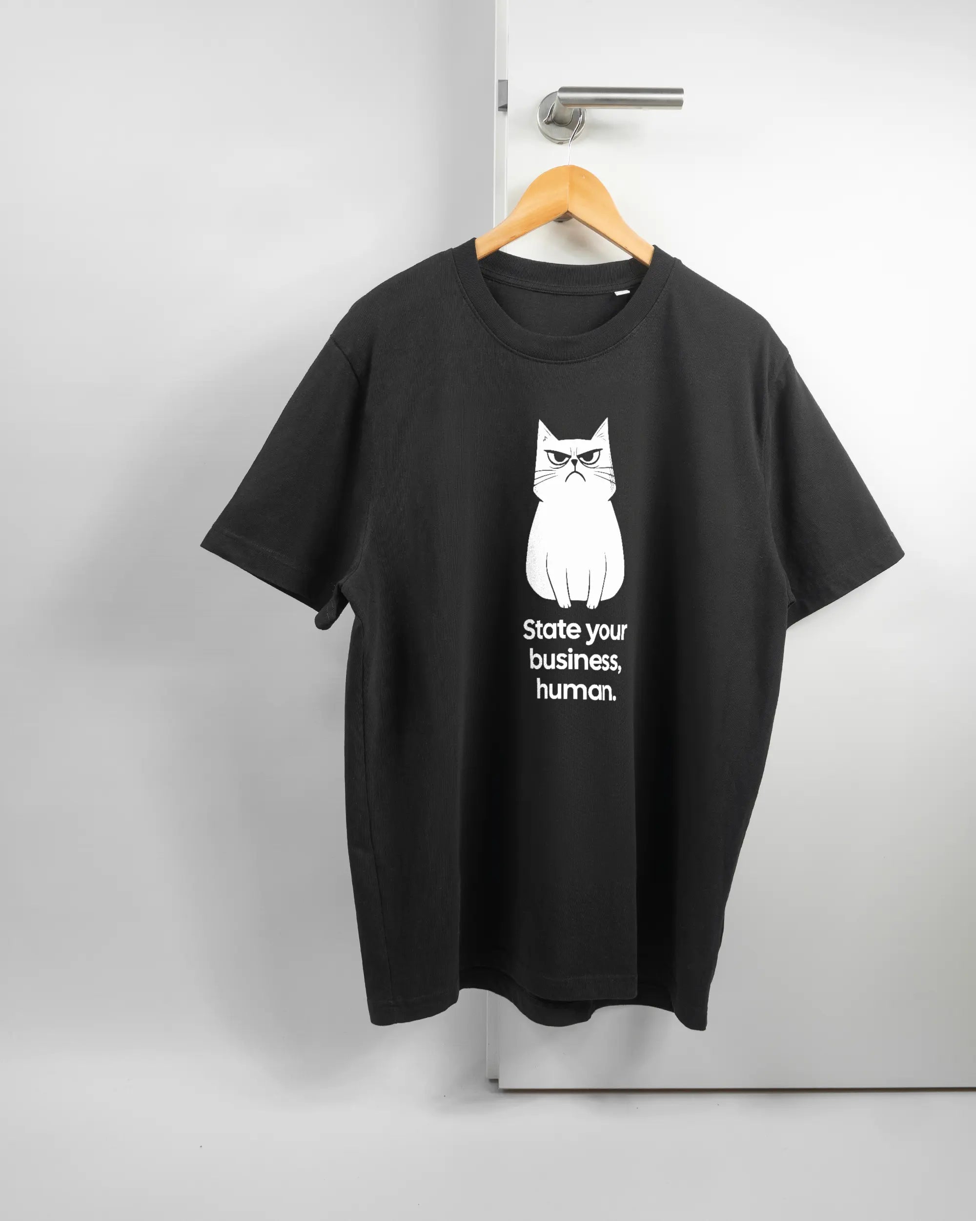 Vorderseite des Organic Unisex T-Shirt Relaxed von Maondo mit dem Design Grumpy Purr in der Farbe Black und in der Größe L auf einem Bügel an der Türklinke aufgehängt