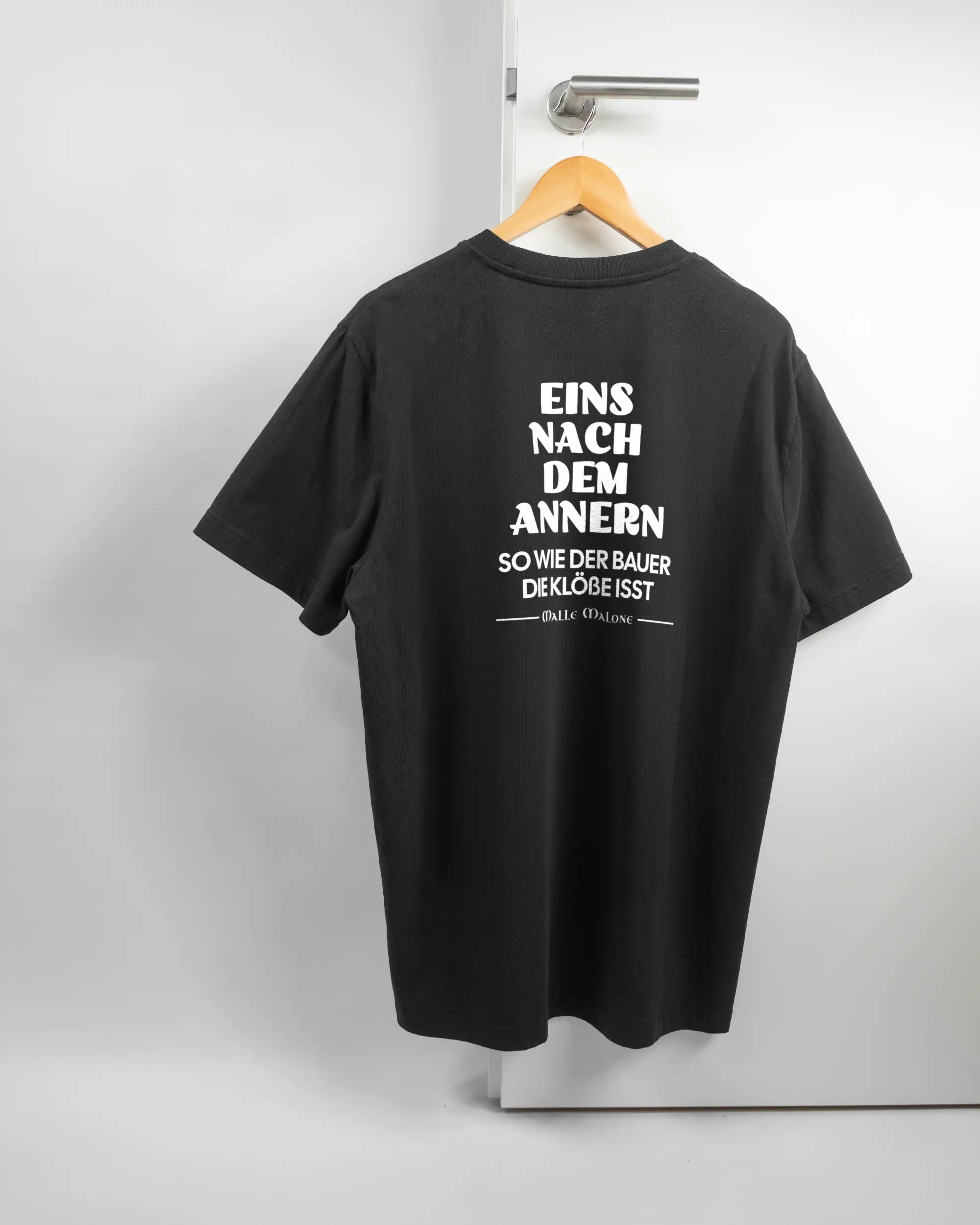 Rückseite des Organic Unisex T-Shirt Relaxed von Maondo mit dem Design Eins nach dem Annern in der Farbe Black und in der Größe L auf einem Bügel an der Türklinke aufgehängt