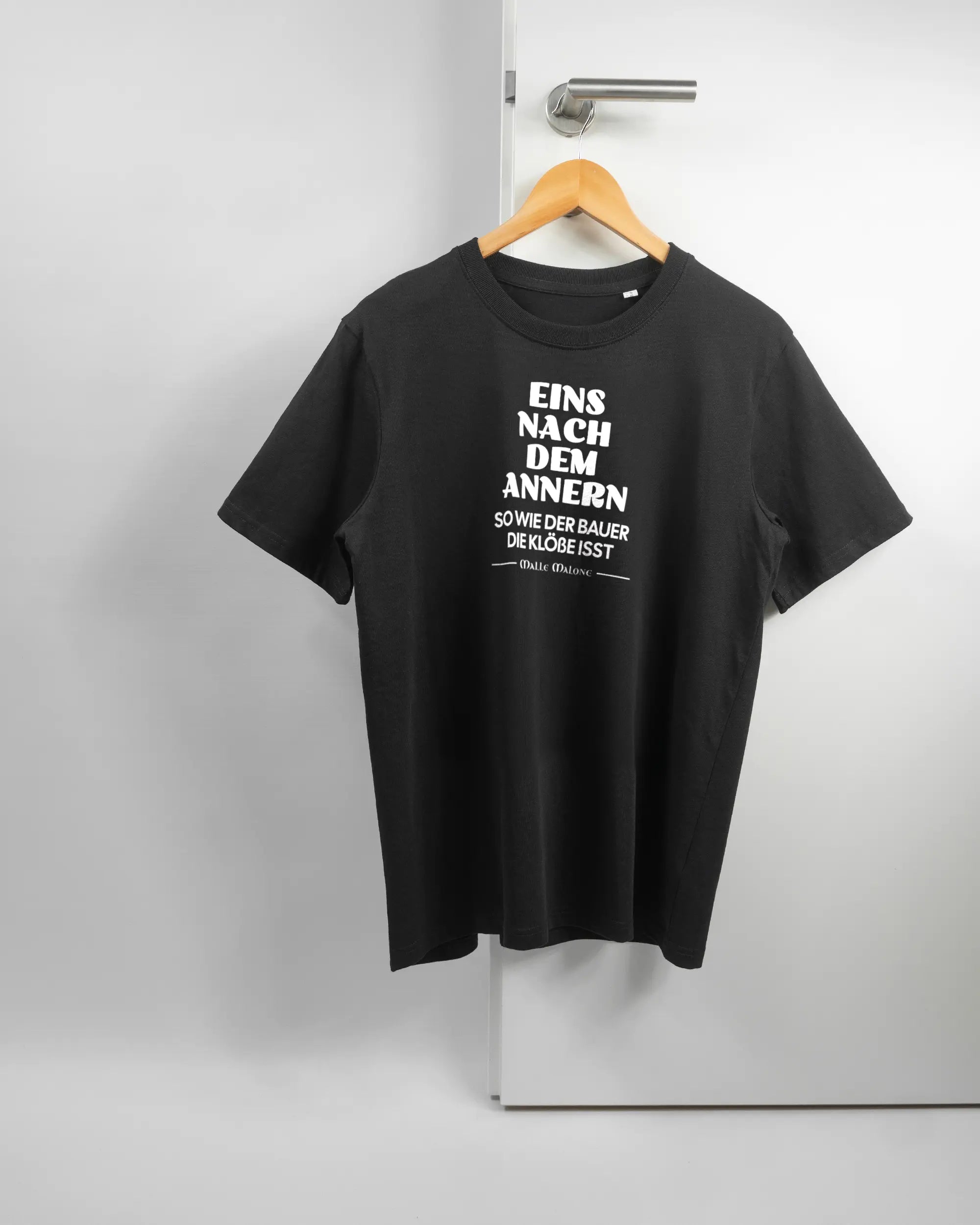 Vorderseite des Organic Unisex T-Shirt Relaxed von Maondo mit dem Design Eins nach dem Annern in der Farbe Black und in der Größe S auf einem Bügel an der Türklinke aufgehängt