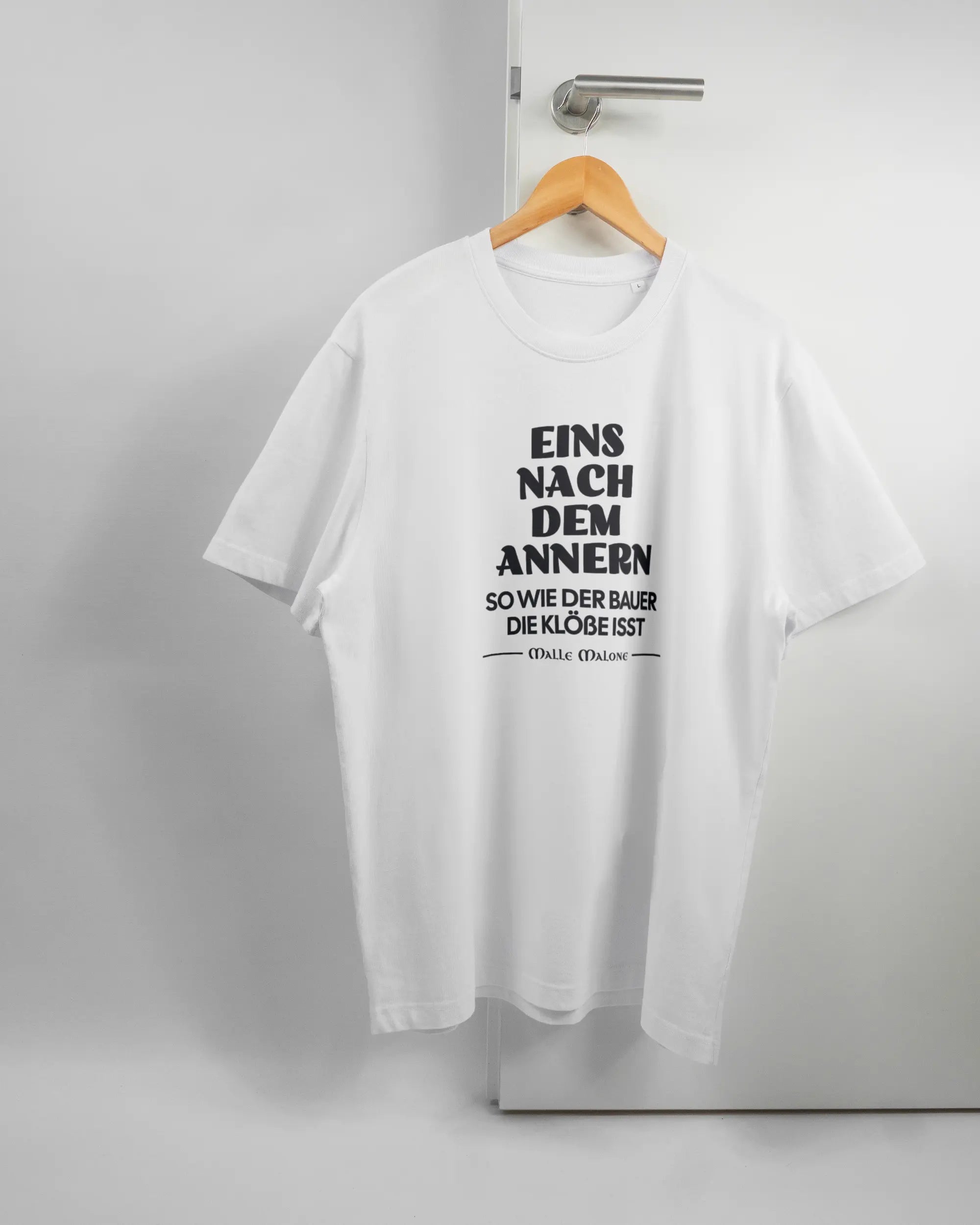 Vorderseite des Organic Unisex T-Shirt Relaxed von Maondo mit dem Design Eins nach dem Annern in der Farbe White und in der Größe L auf einem Bügel an der Türklinke aufgehängt