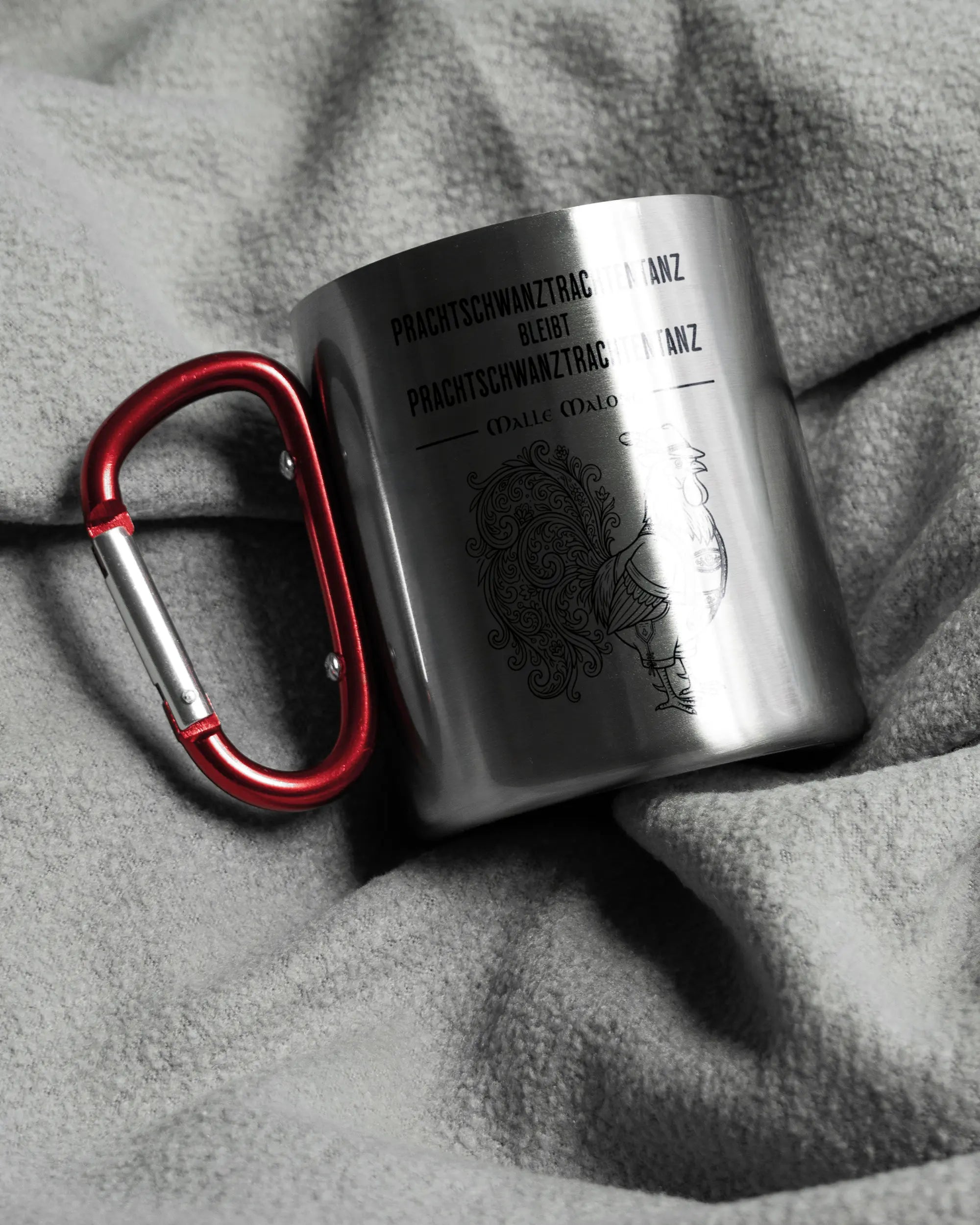 Aufnahme, die die linke Seite der beidseitig bedruckten Edelstahltasse von Maondo auf einer flauschigen Fließdecke mit dem Design Trachtentanz in der Farbe Metallic-Red zeigt