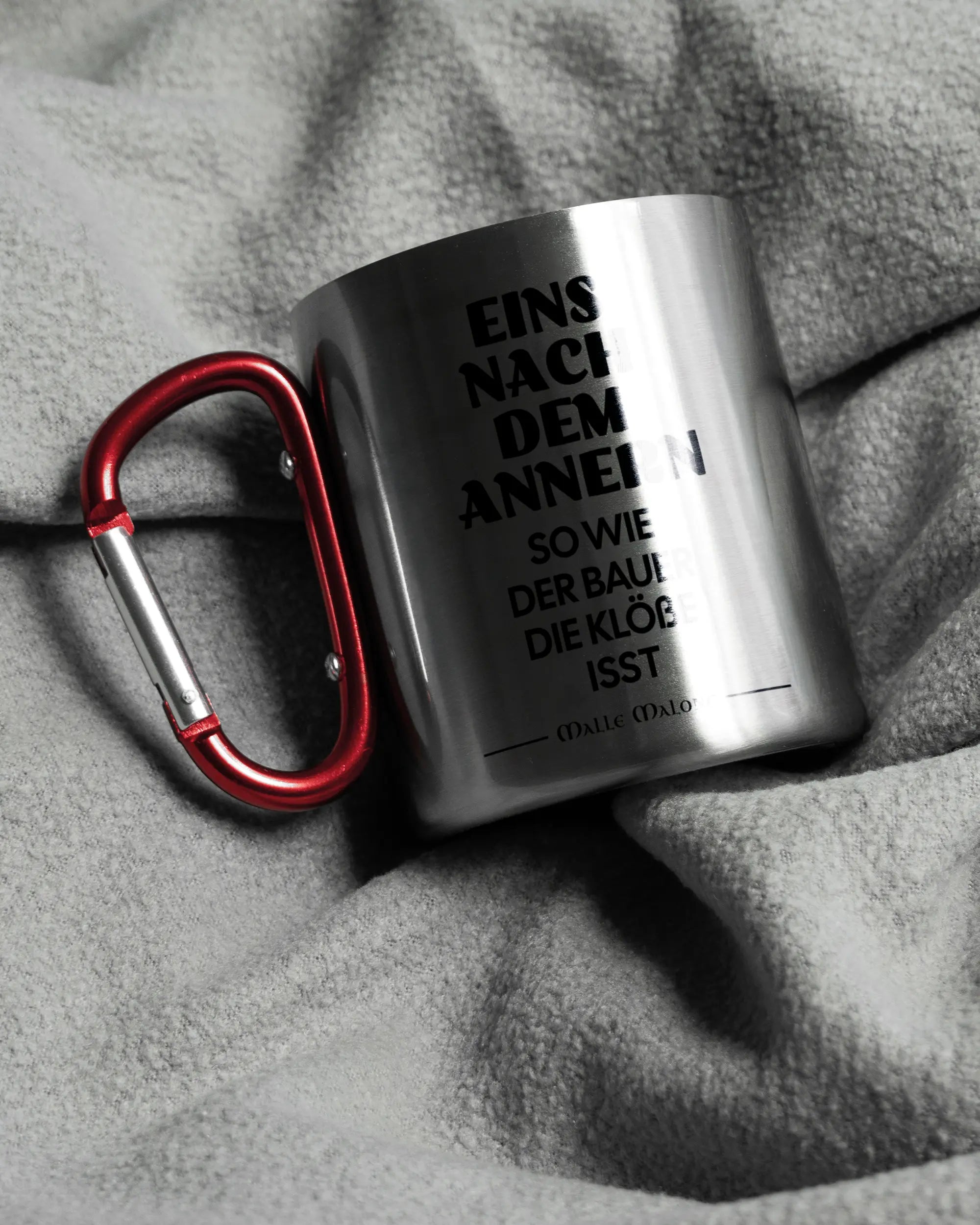 Aufnahme, die die linke Seite der beidseitig bedruckten Edelstahltasse von Maondo auf einer flauschigen Fließdecke mit dem Design Eins nach dem Annern in der Farbe Metallic-Red zeigt