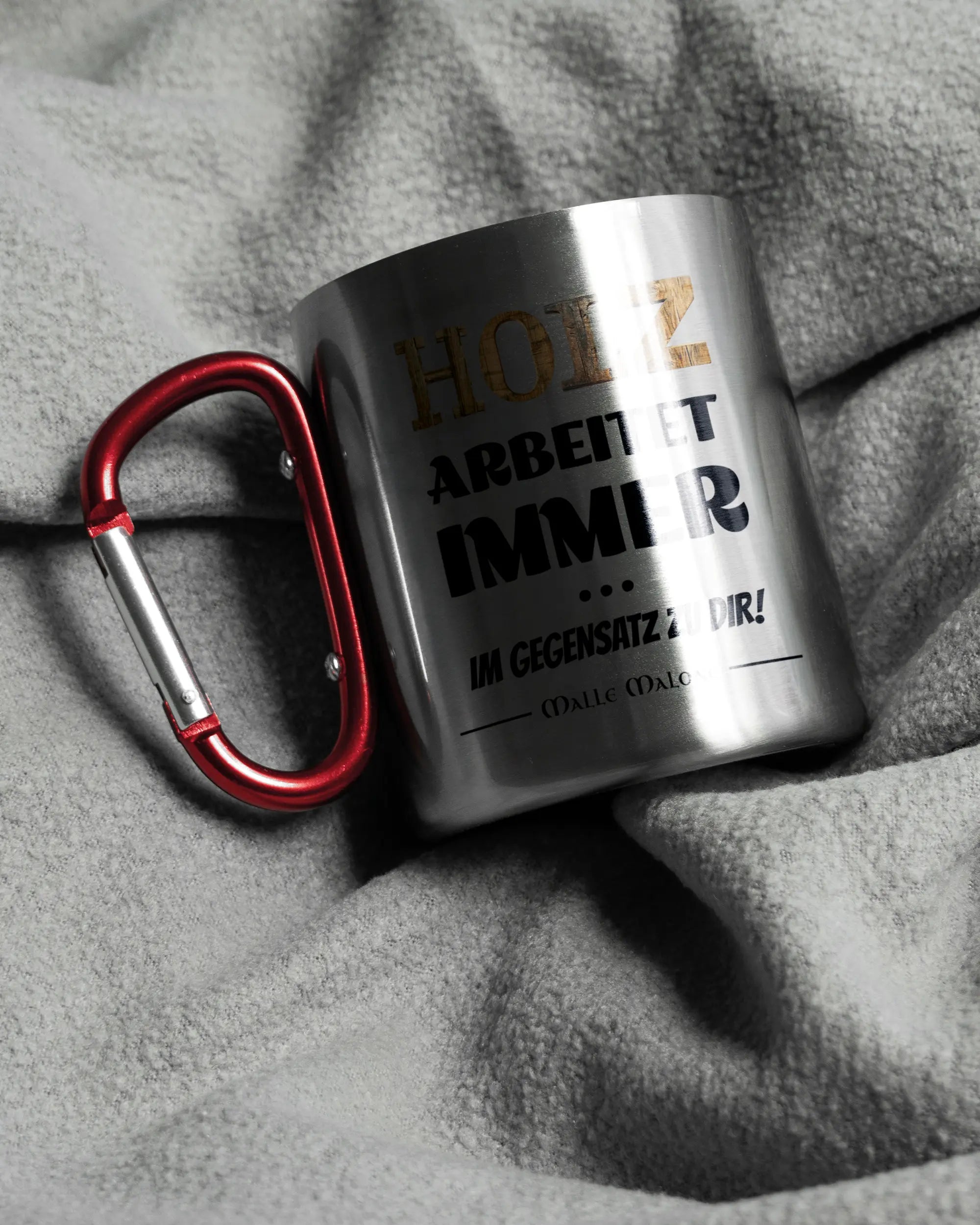 Aufnahme, die die linke Seite der beidseitig bedruckten Edelstahltasse von Maondo auf einer flauschigen Fließdecke mit dem Design Holzarbeit in der Farbe Metallic-Red zeigt