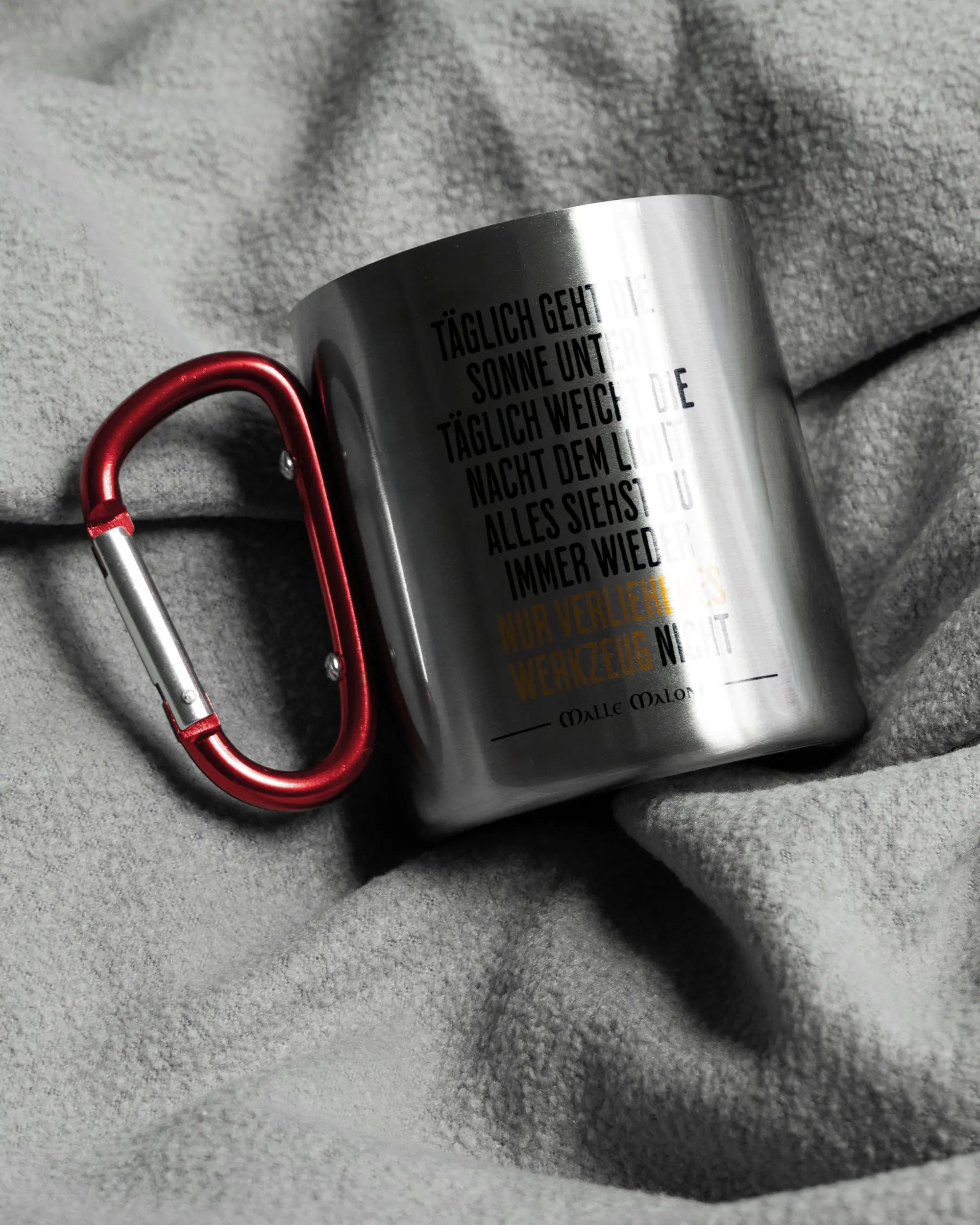 Aufnahme, die die linke Seite der beidseitig bedruckten Edelstahltasse von Maondo auf einer flauschigen Fließdecke mit dem Design Wegzeug in der Farbe Metallic-Red zeigt