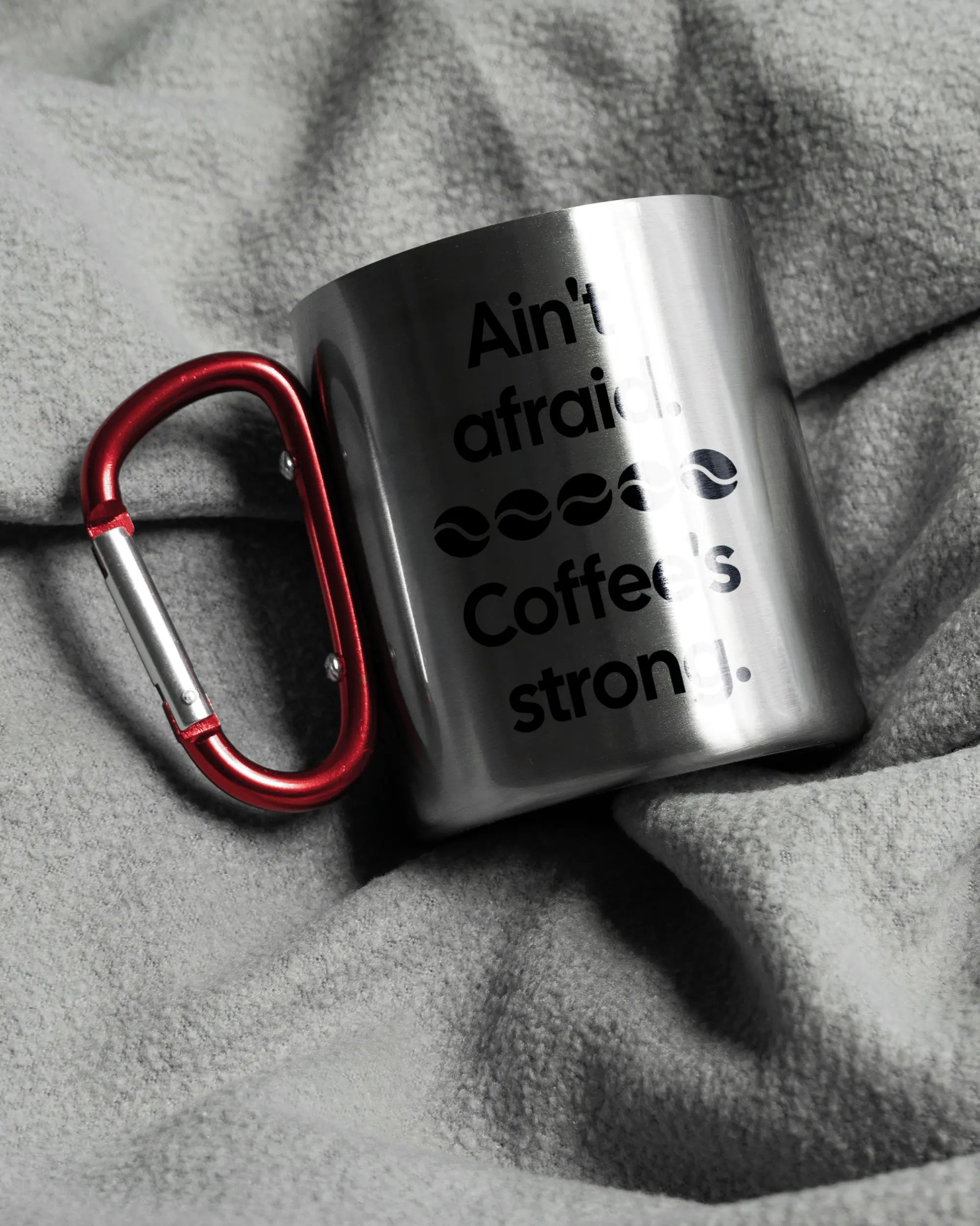 Aufnahme, die die linke Seite der beidseitig bedruckten Edelstahltasse von Maondo auf einer flauschigen Fließdecke mit dem Design Coffee is strong in der Farbe Metallic-Red zeigt