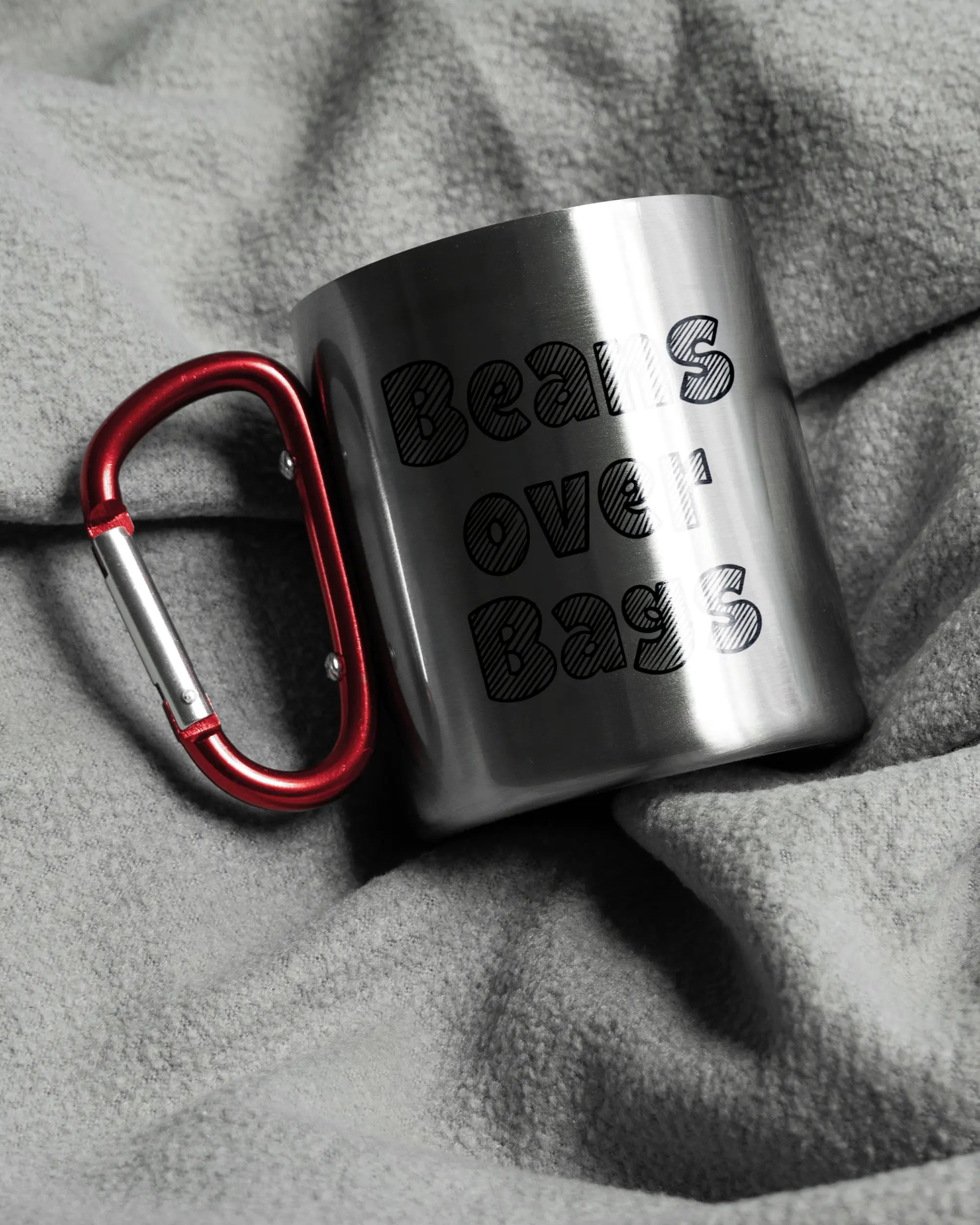 Aufnahme, die die linke Seite der beidseitig bedruckten Edelstahltasse von Maondo auf einer flauschigen Fließdecke mit dem Design Beans over Bags in der Farbe Metallic-Red zeigt