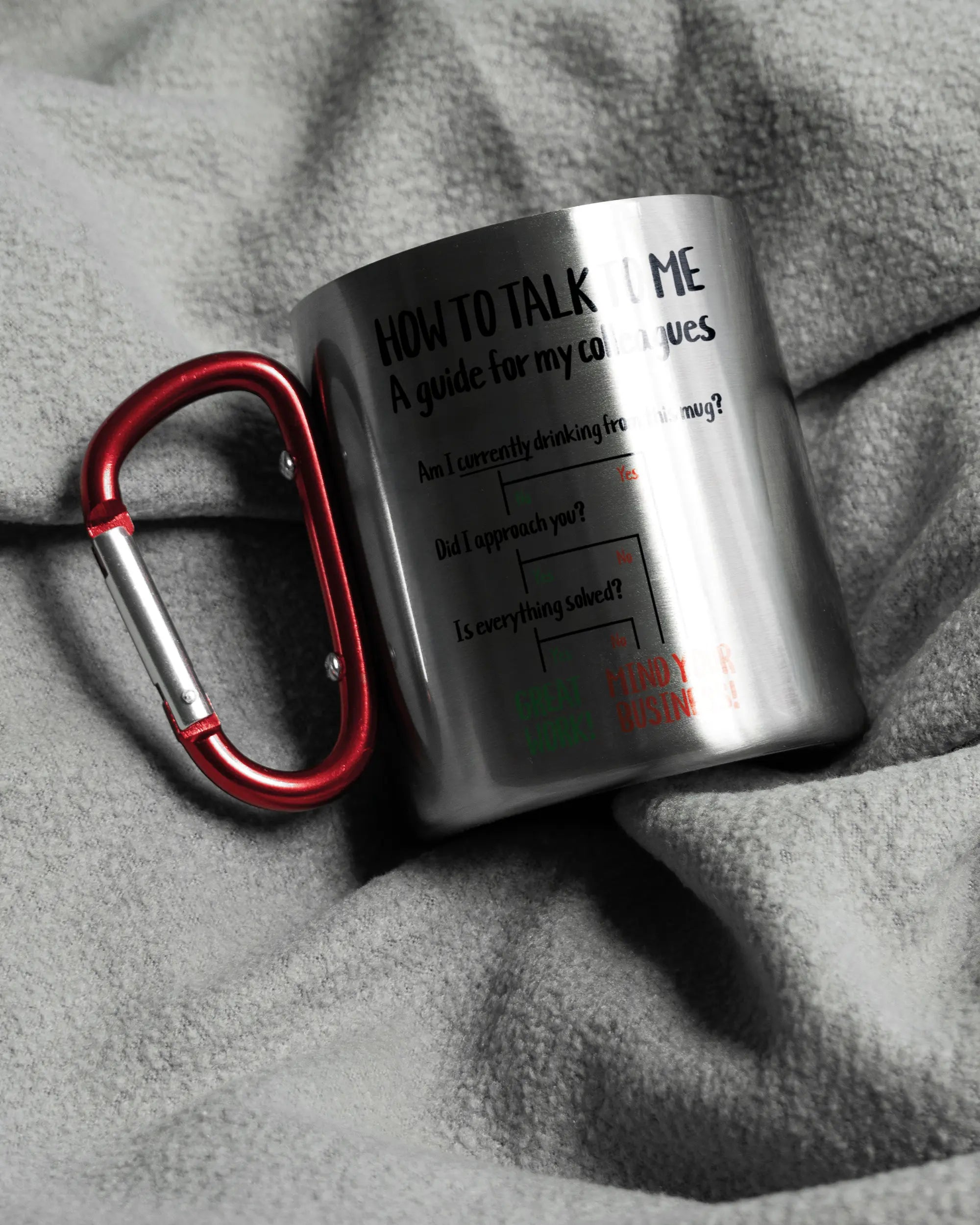 Aufnahme, die die linke Seite der beidseitig bedruckten Edelstahltasse von Maondo auf einer flauschigen Fließdecke mit dem Design Colleagues Guide in der Farbe Metallic-Red zeigt