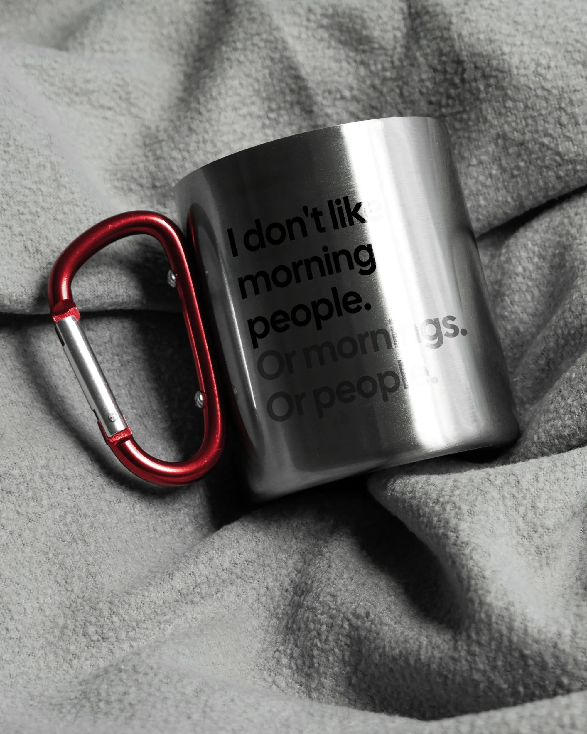 Aufnahme, die die linke Seite der beidseitig bedruckten Edelstahltasse von Maondo auf einer flauschigen Fließdecke mit dem Design Morning Person in der Farbe Metallic-Red zeigt