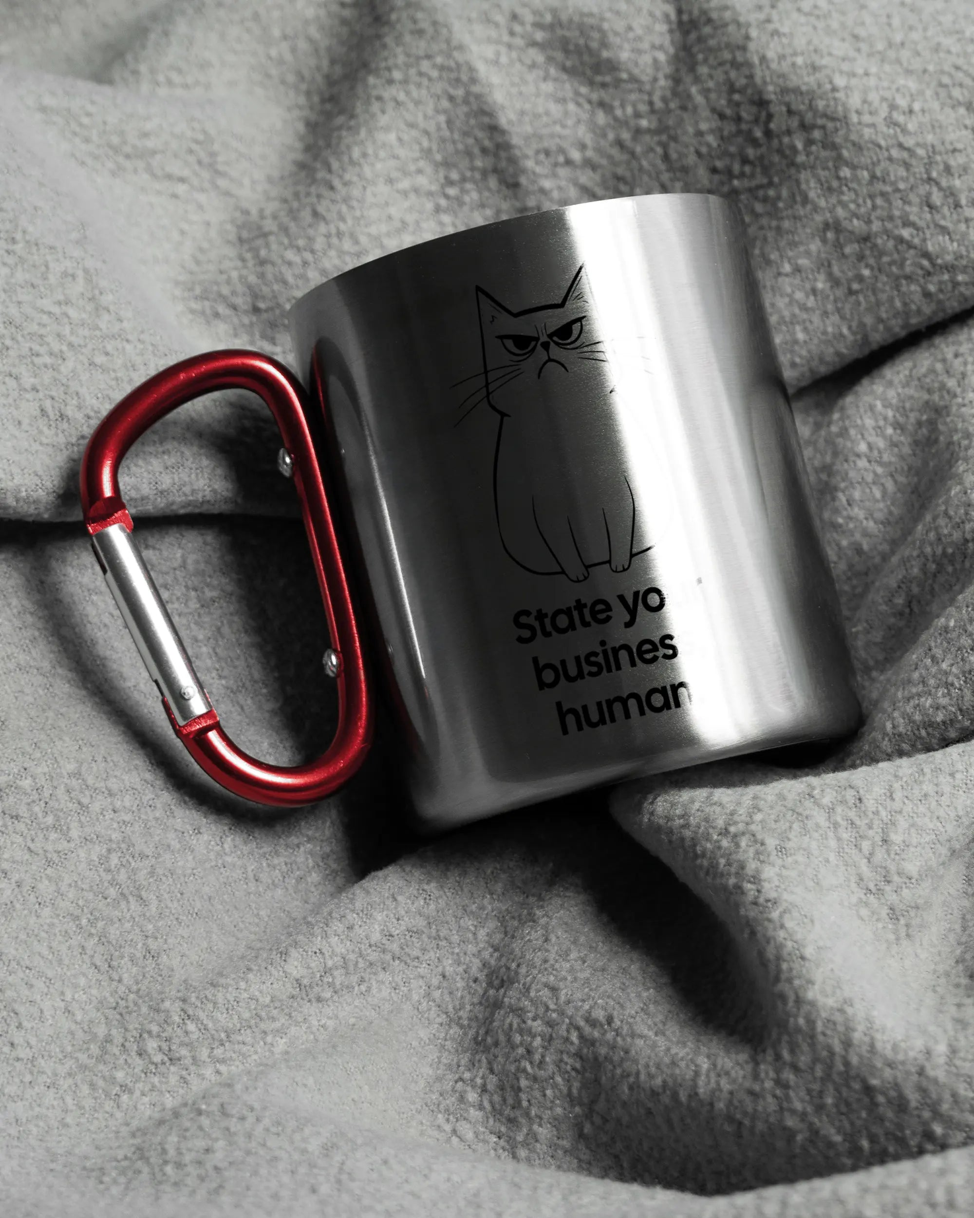 Aufnahme, die die linke Seite der beidseitig bedruckten Edelstahltasse von Maondo auf einer flauschigen Fließdecke mit dem Design Grumpy Purr in der Farbe Metallic-Red zeigt