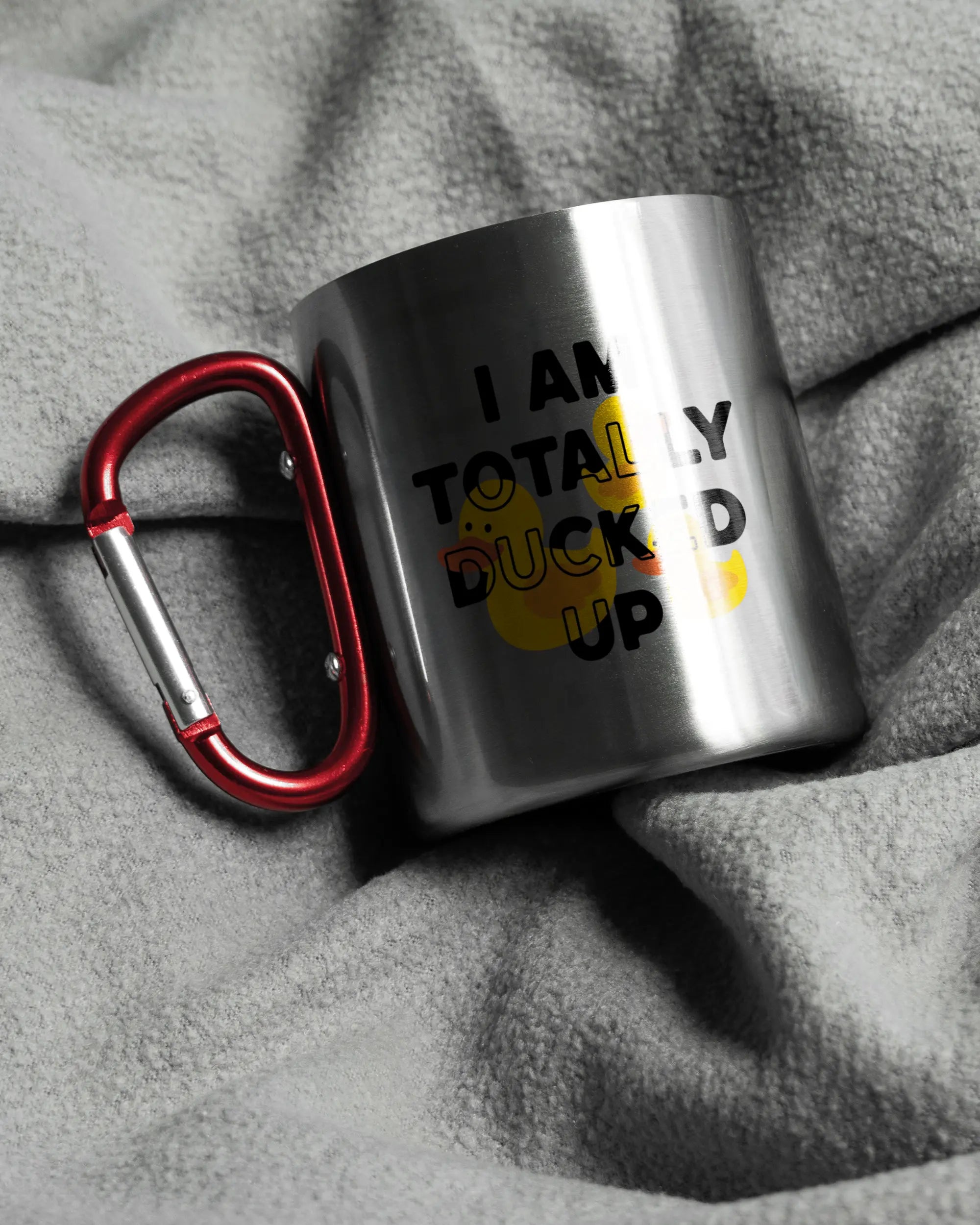 Aufnahme, die die linke Seite der beidseitig bedruckten Edelstahltasse von Maondo auf einer flauschigen Fließdecke mit dem Design Ducked Up in der Farbe Metallic-Red zeigt