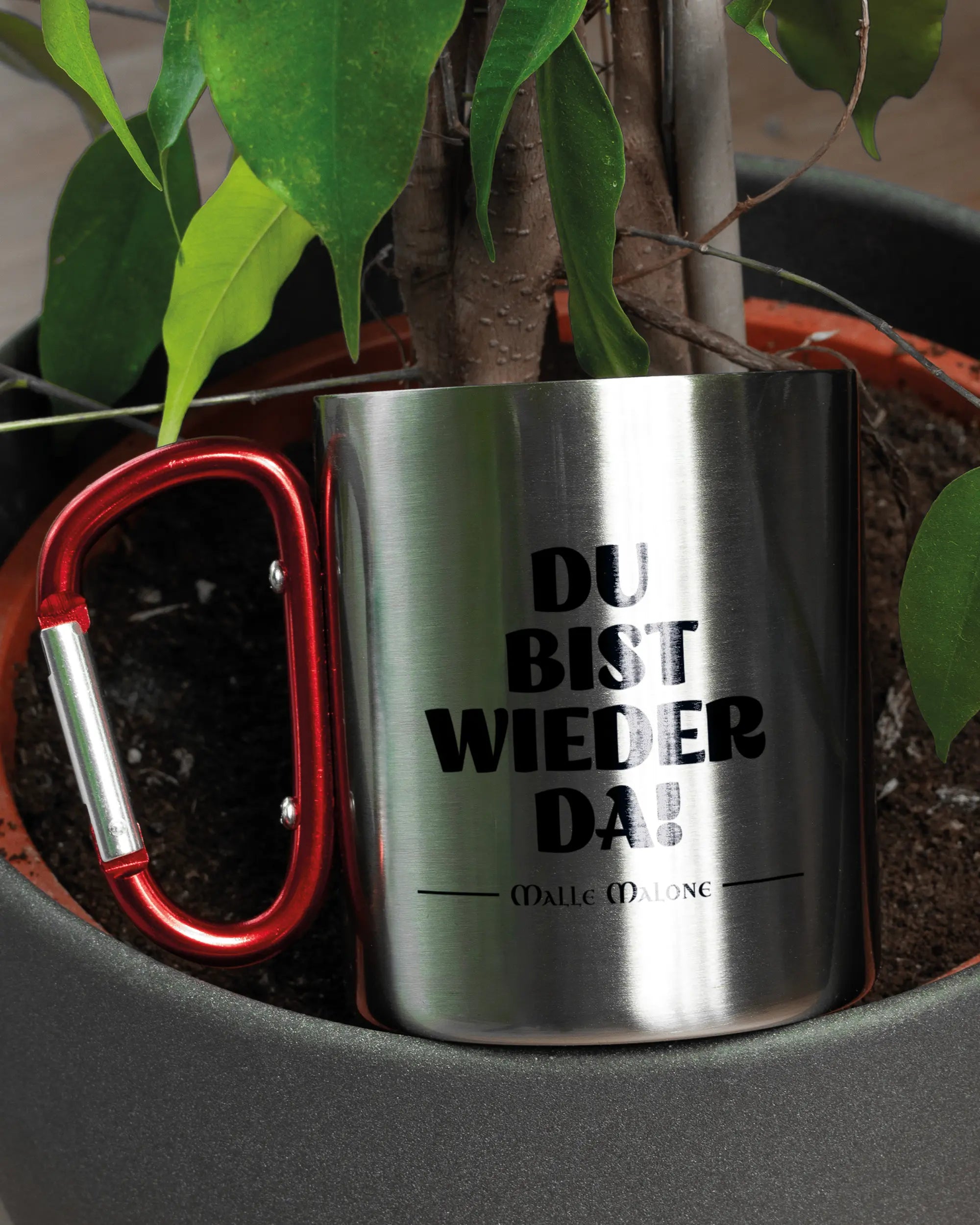 Aufnahme der linken Seite der beidseitig bedruckten Edelstahltasse von Maondo mit dem Design Wieder da in der Farbe Metallic-Red in einem Blumentopf
