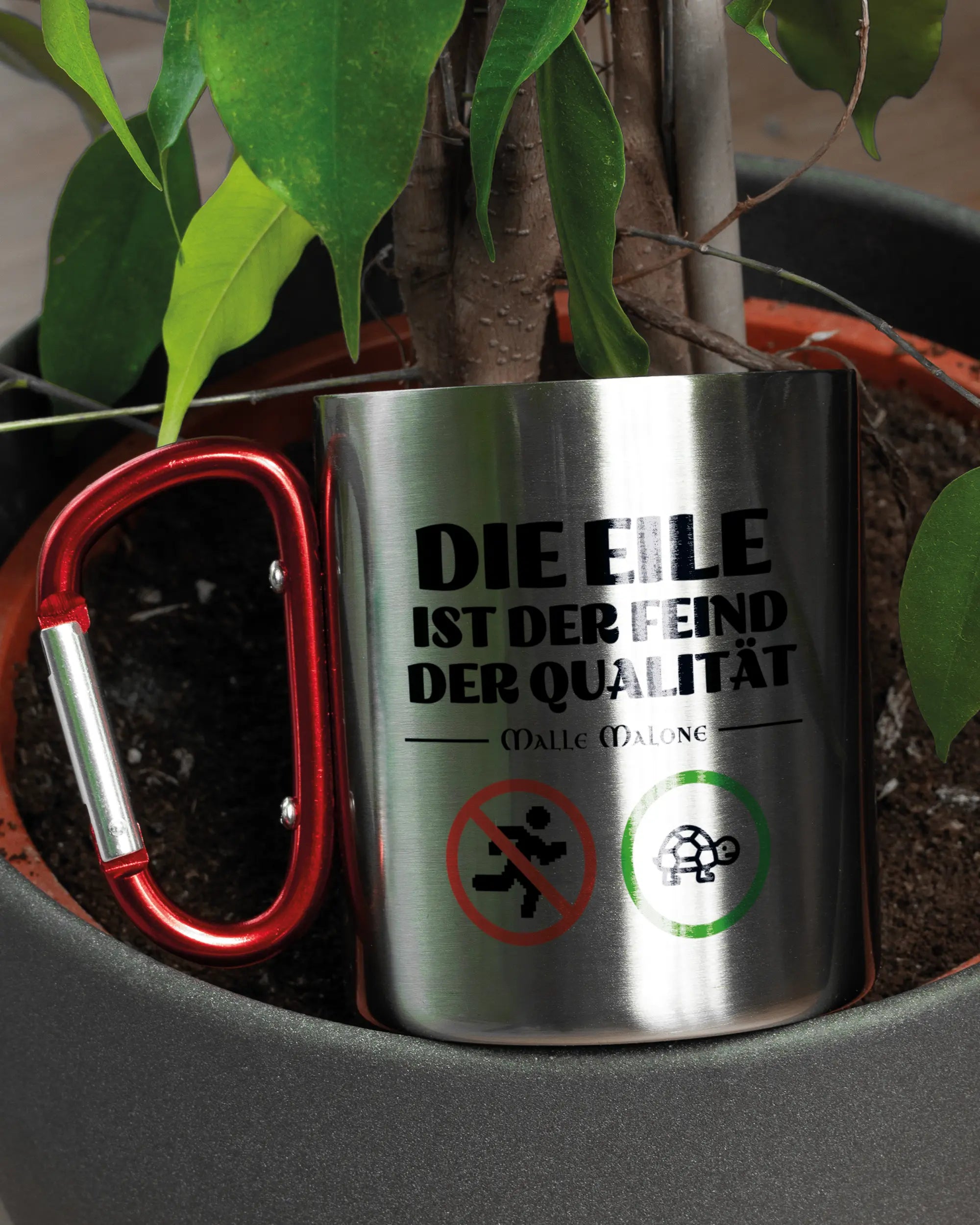 Aufnahme der linken Seite der beidseitig bedruckten Edelstahltasse von Maondo mit dem Design Keine Eile in der Farbe Metallic-Red in einem Blumentopf