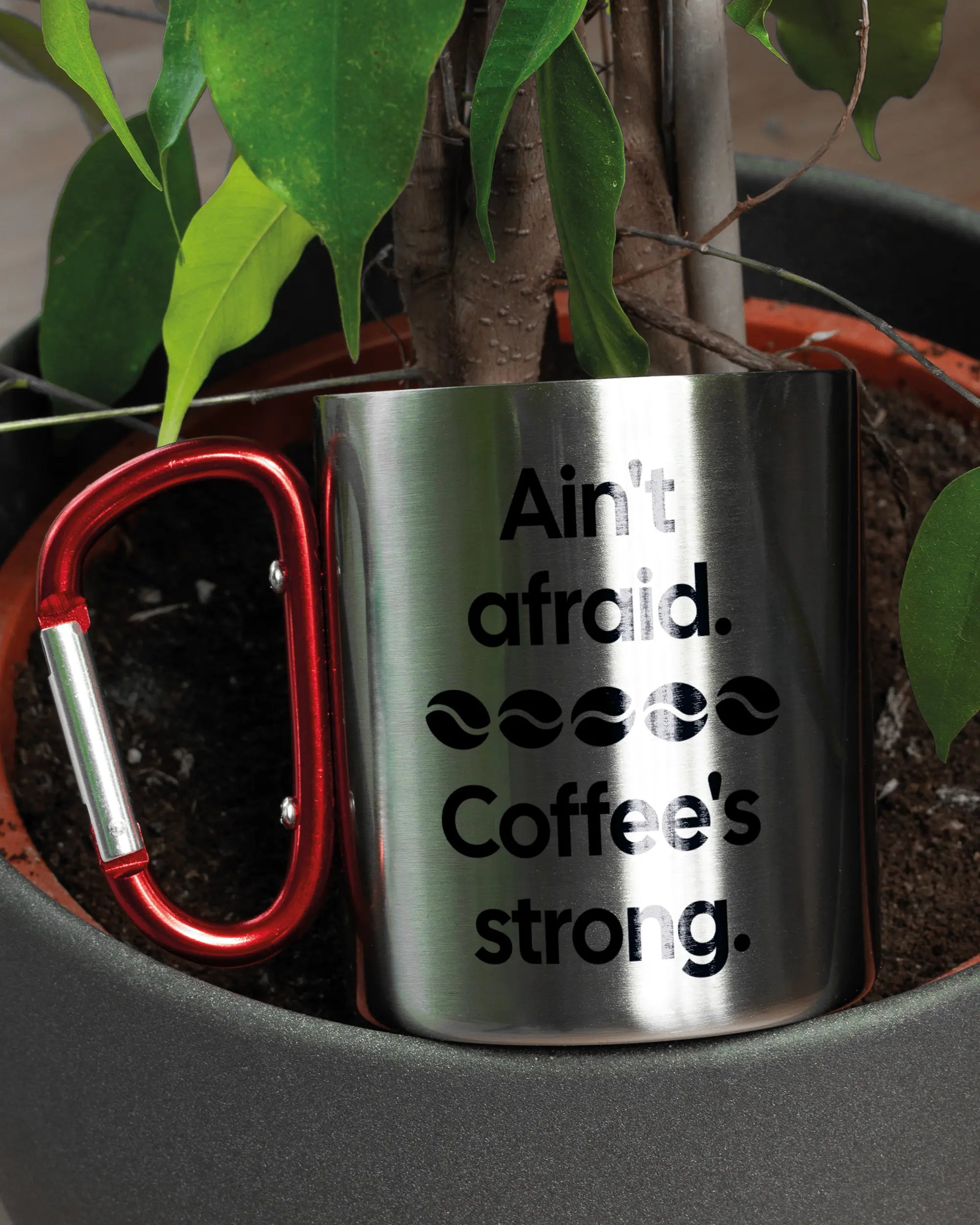 Aufnahme der linken Seite der beidseitig bedruckten Edelstahltasse von Maondo mit dem Design Coffee is strong in der Farbe Metallic-Red in einem Blumentopf
