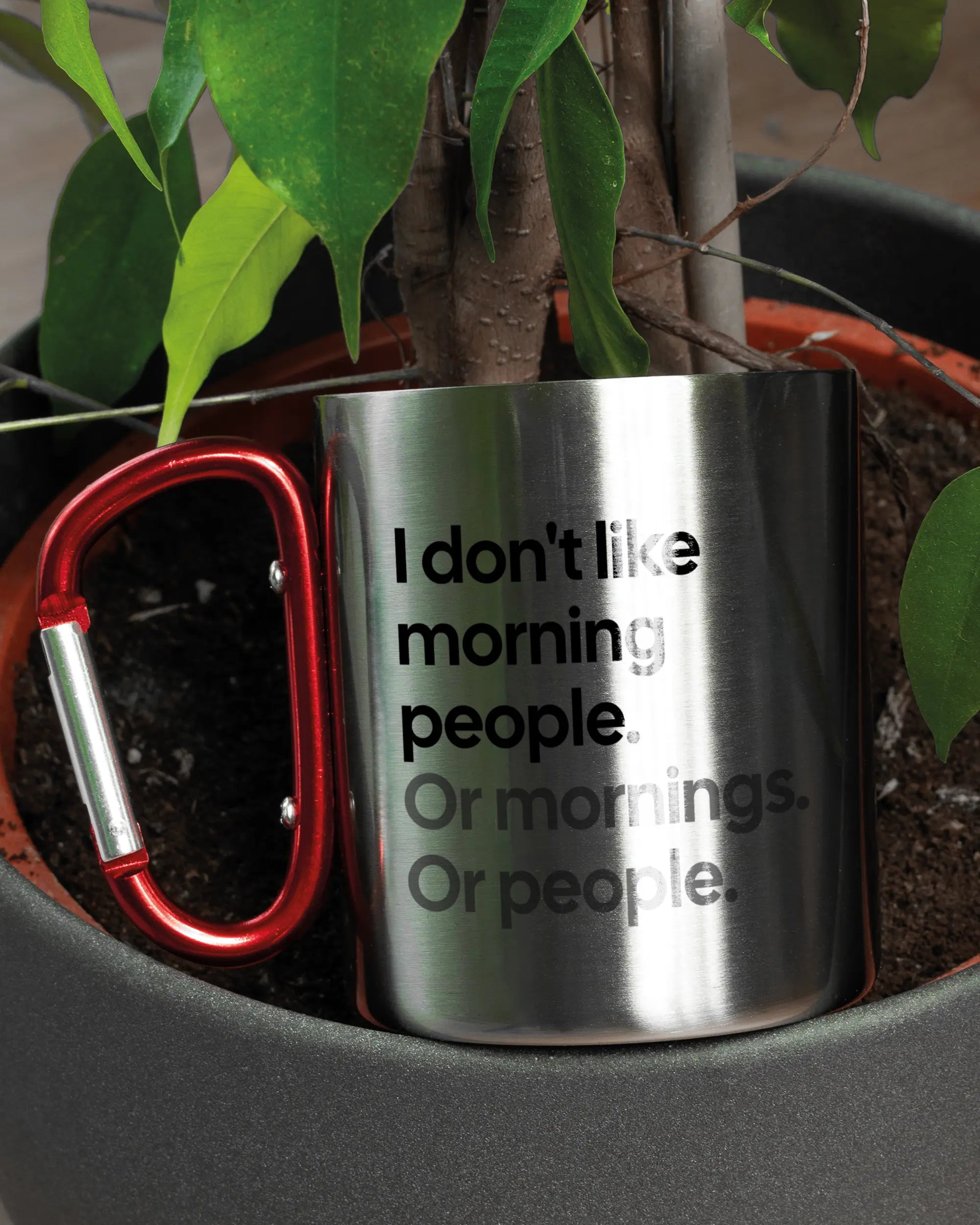 Aufnahme der linken Seite der beidseitig bedruckten Edelstahltasse von Maondo mit dem Design Morning Person in der Farbe Metallic-Red in einem Blumentopf