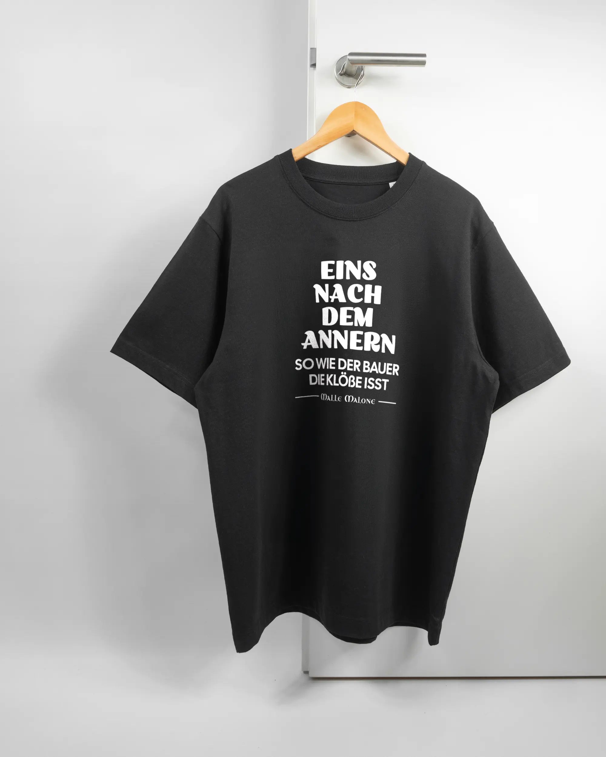 Vorderseite des Organic Unisex T-Shirt Heavy Oversized von Maondo mit dem Design Eins nach dem Annern in der Farbe Black und in der Größe L auf einem Bügel an der Türklinke aufgehängt