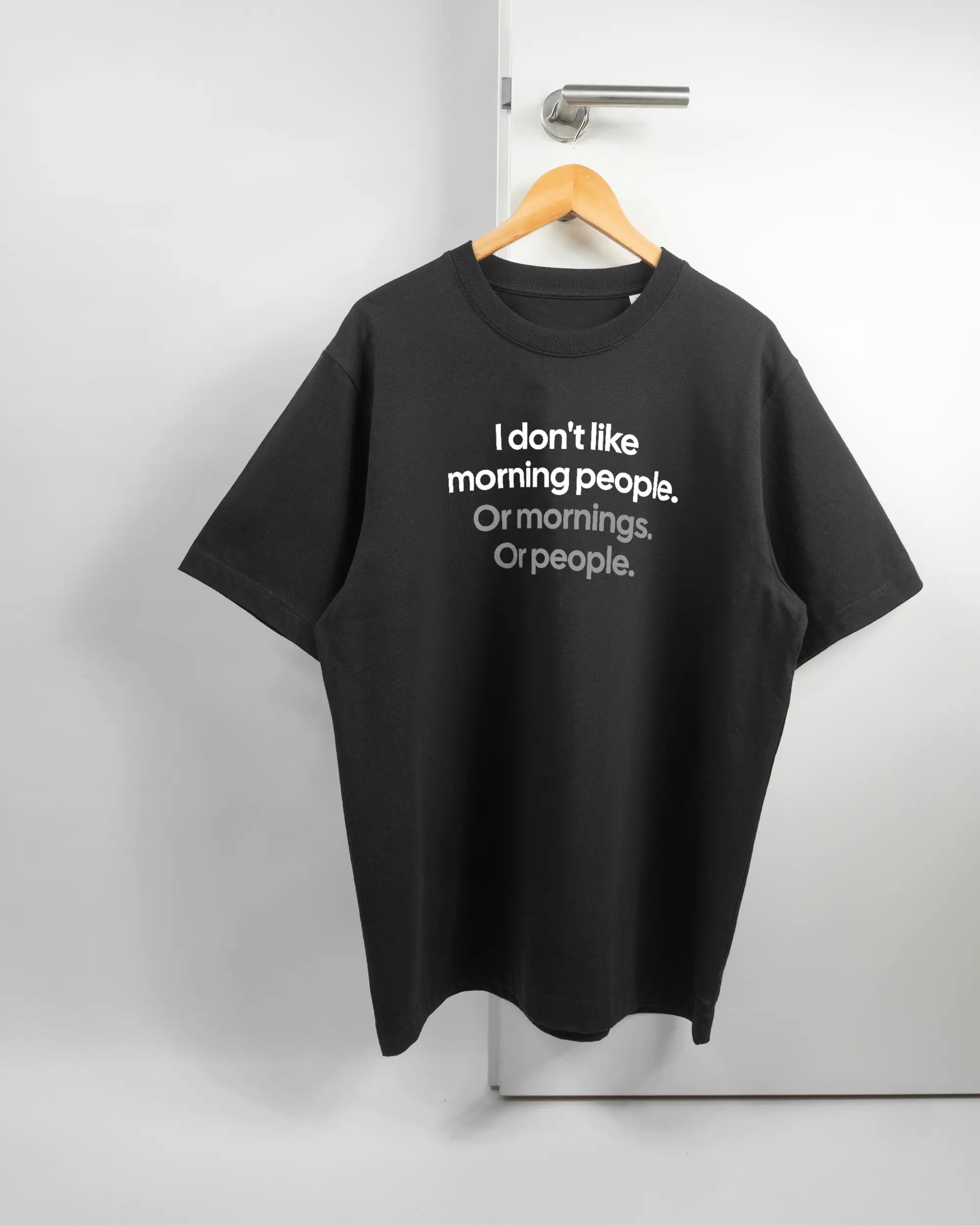 Vorderseite des Organic Unisex T-Shirt Heavy Oversized von Maondo mit dem Design Morning Person in der Farbe Black und in der Größe L auf einem Bügel an der Türklinke aufgehängt