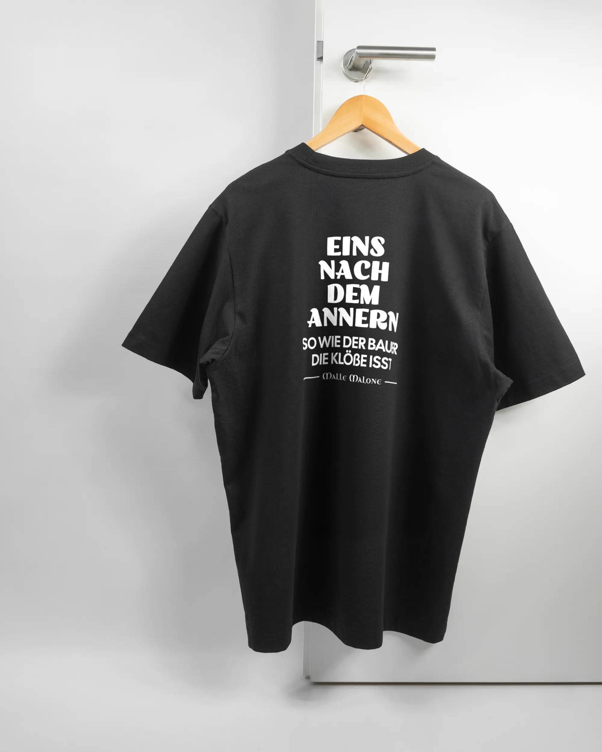 Rückseite des Organic Unisex T-Shirt Heavy Oversized von Maondo mit dem Design Eins nach dem Annern in der Farbe Black und in der Größe L auf einem Bügel an der Türklinke aufgehängt