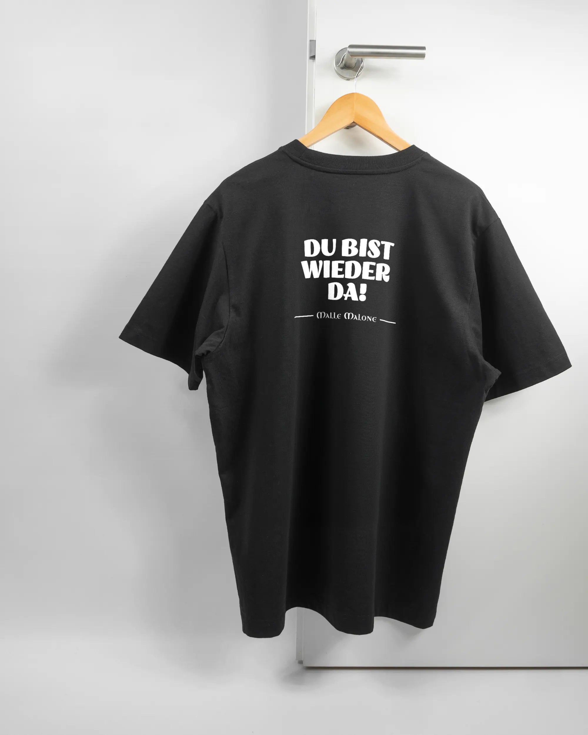 Rückseite des Organic Unisex T-Shirt Heavy Oversized von Maondo mit dem Design Wieder da in der Farbe Black und in der Größe L auf einem Bügel an der Türklinke aufgehängt