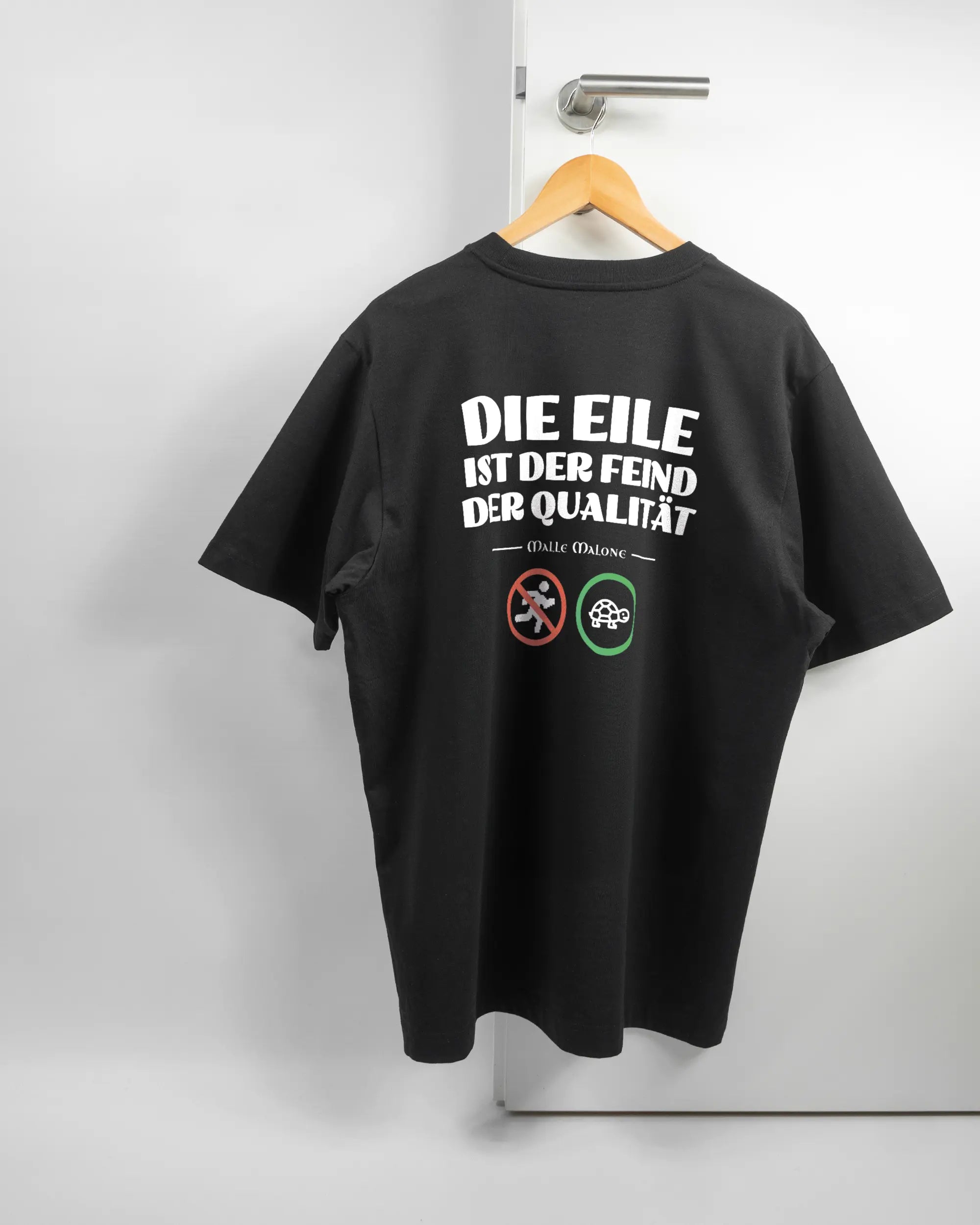 Rückseite des Organic Unisex T-Shirt Heavy Oversized von Maondo mit dem Design Keine Eile in der Farbe Black und in der Größe L auf einem Bügel an der Türklinke aufgehängt