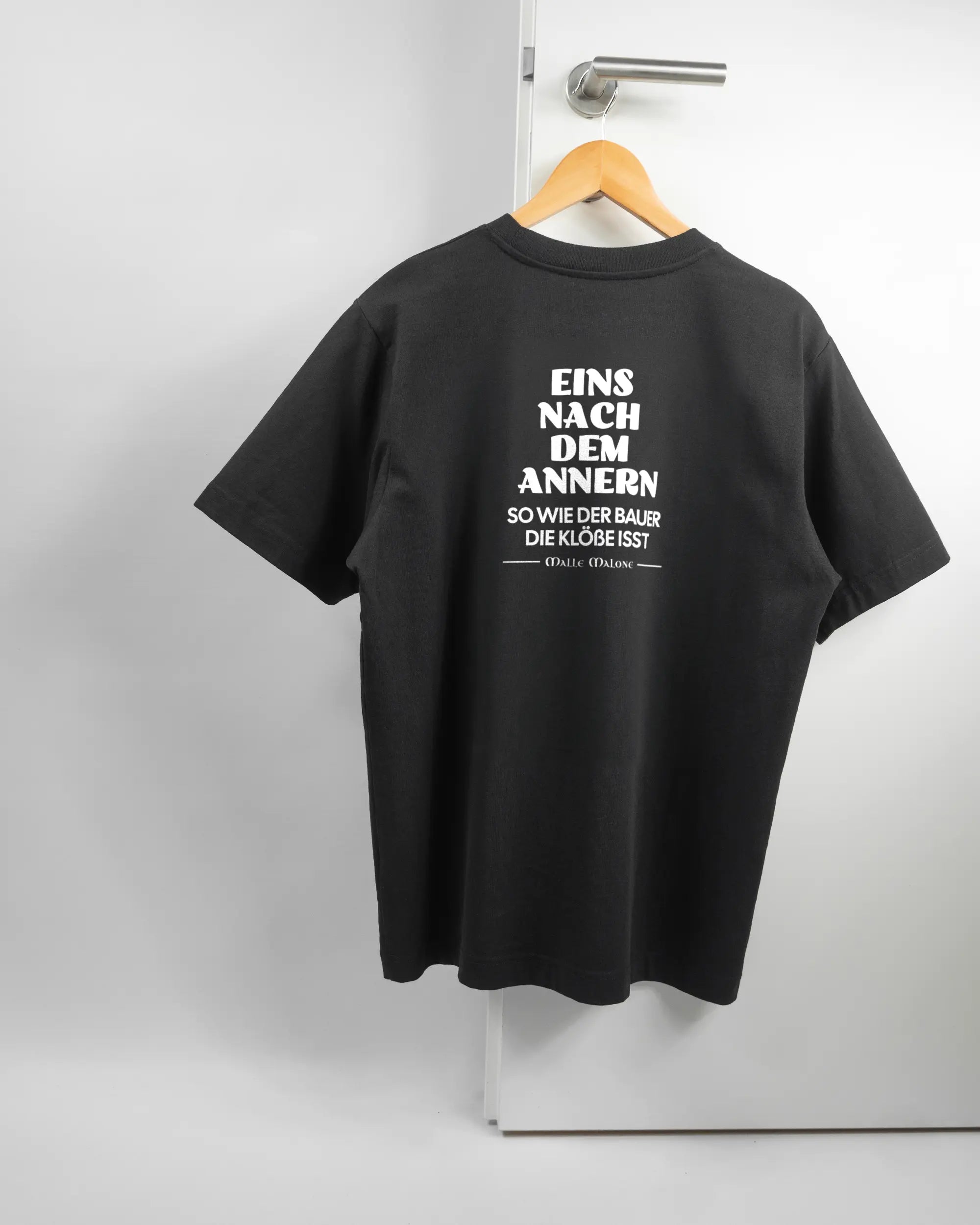 Rückseite des Organic Unisex T-Shirt Heavy Oversized von Maondo mit dem Design Eins nach dem Annern in der Farbe Black und in der Größe S auf einem Bügel an der Türklinke aufgehängt
