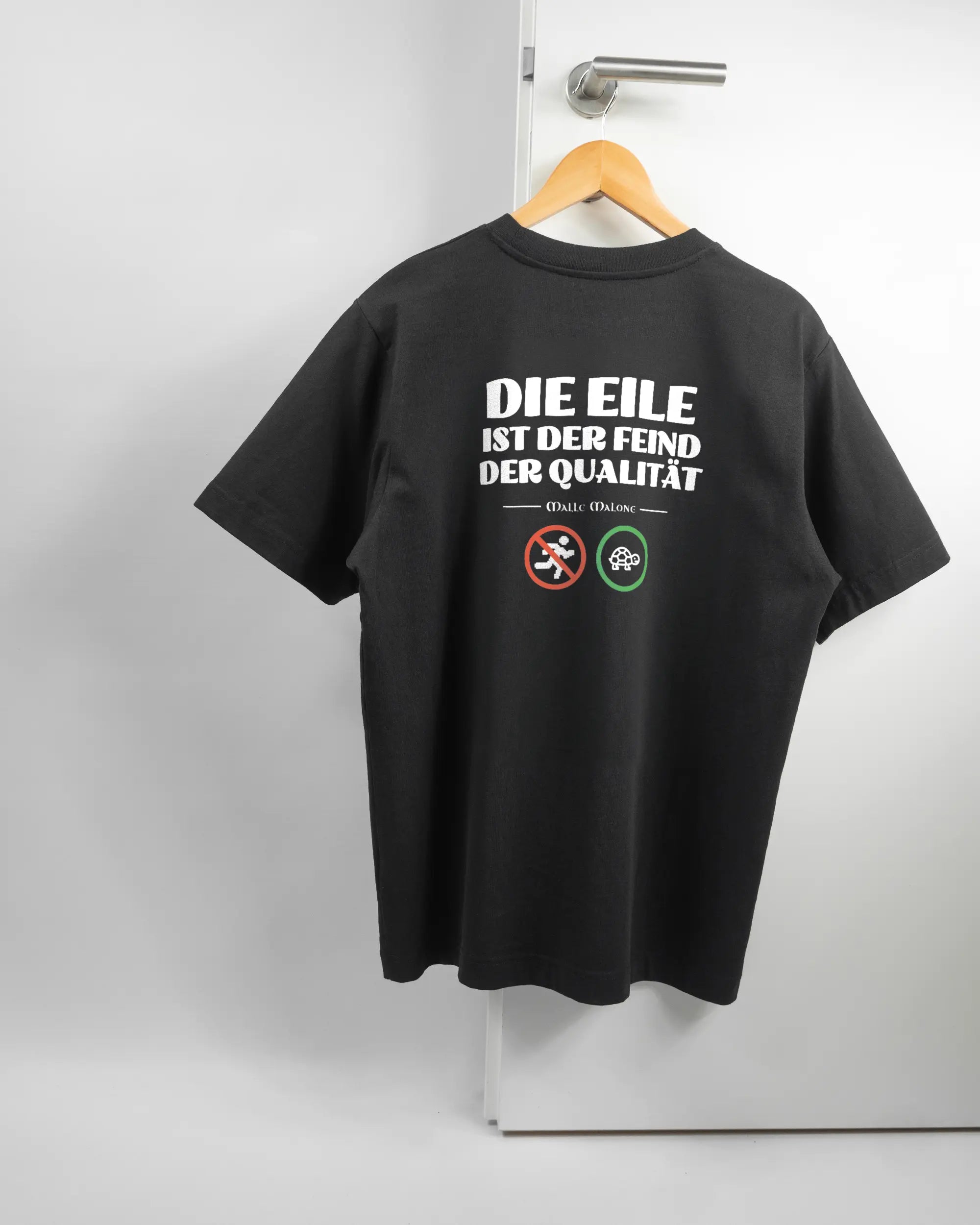 Rückseite des Organic Unisex T-Shirt Heavy Oversized von Maondo mit dem Design Keine Eile in der Farbe Black und in der Größe S auf einem Bügel an der Türklinke aufgehängt