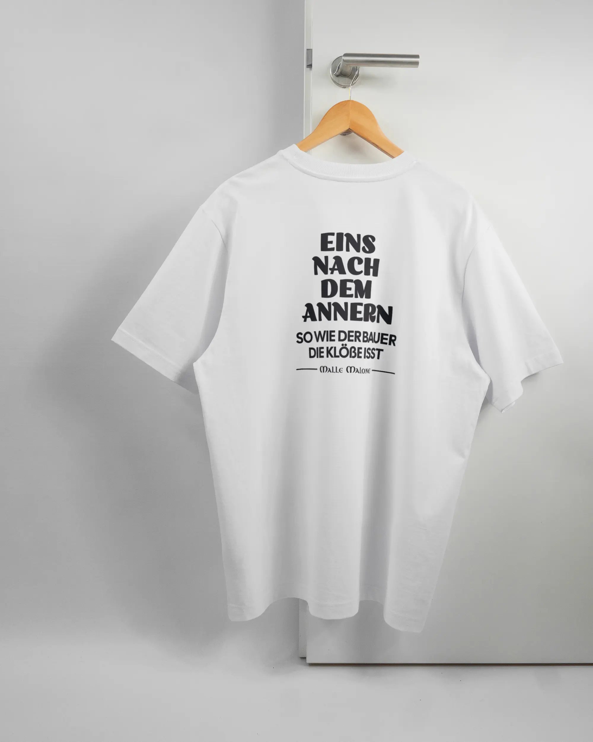 Rückseite des Organic Unisex T-Shirt Heavy Oversized von Maondo mit dem Design Eins nach dem Annern in der Farbe White und in der Größe L auf einem Bügel an der Türklinke aufgehängt