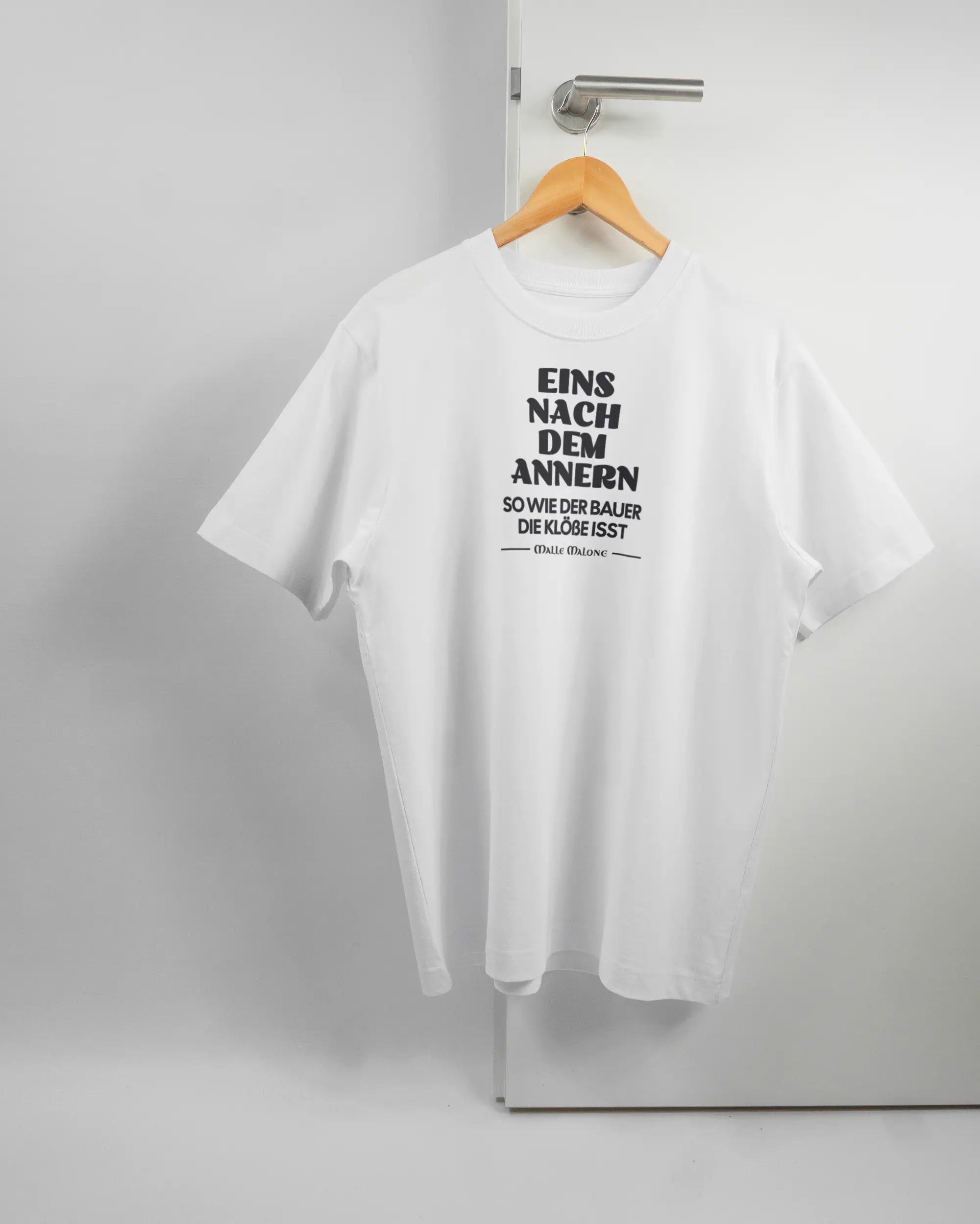 Vorderseite des Organic Unisex T-Shirt Heavy Oversized von Maondo mit dem Design Eins nach dem Annern in der Farbe White und in der Größe S auf einem Bügel an der Türklinke aufgehängt
